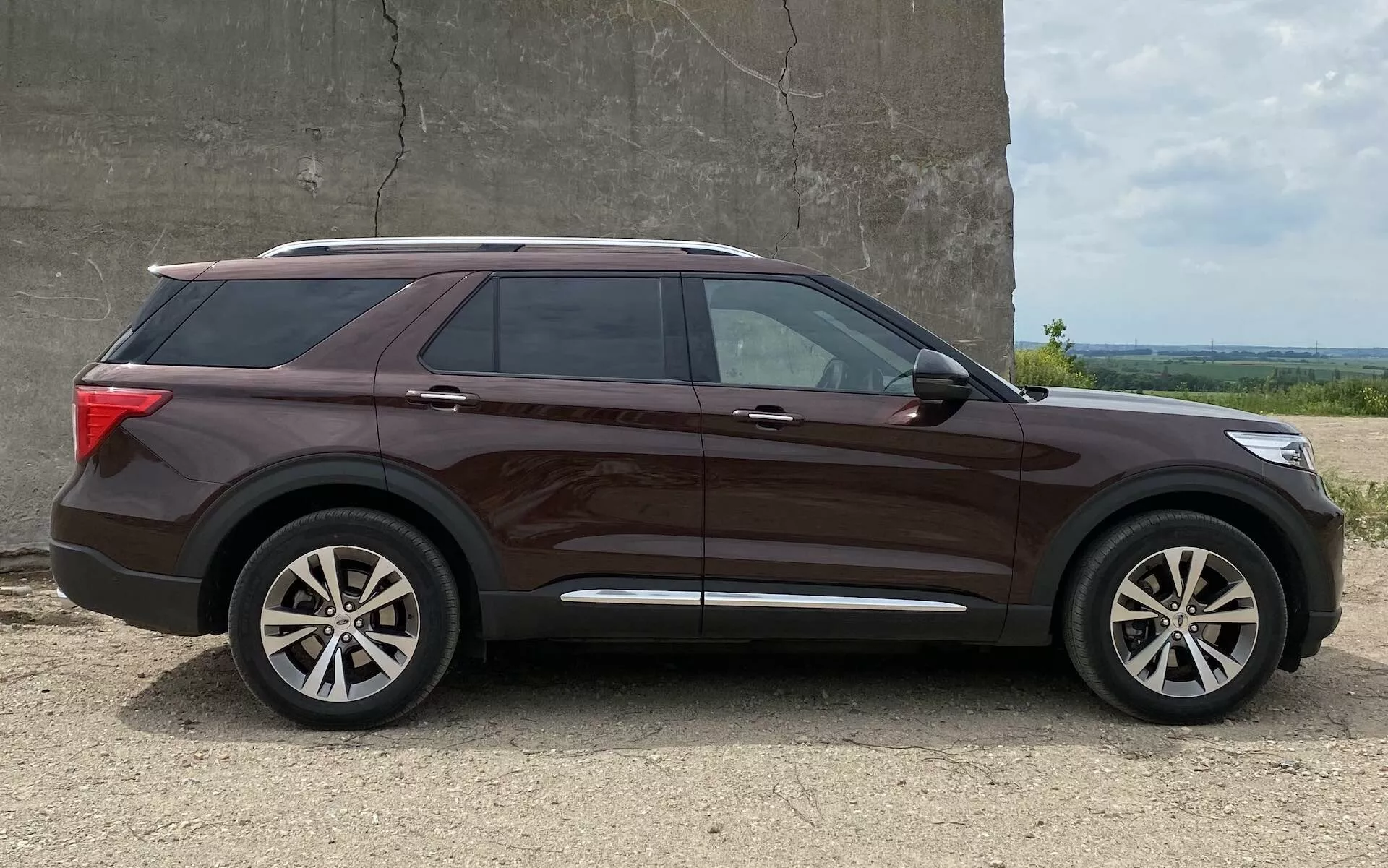Profilvy av en modern medelstor SUV, 2019 års Ford Explorer, med metallisk-brun lackfinish, flerspokes aluminiumfälgar, tonade rutor och takräcken. Fordonet är parkerat på en grusyta med en slät vägg och öppen himmel i bakgrunden, fångat i ljus mitt på dagen.