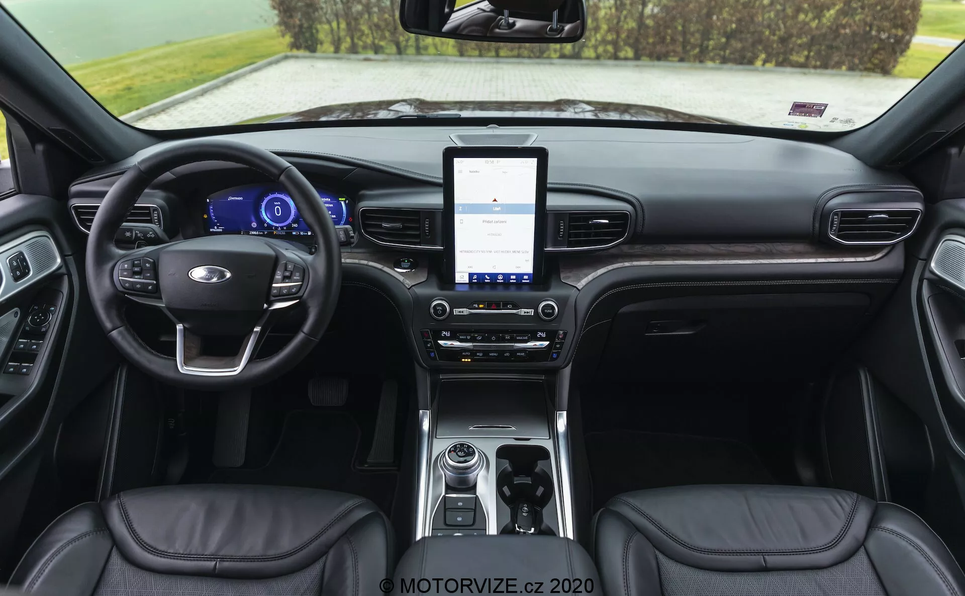 Interiör av en 2019 Ford Explorer som visar instrumentbrädan från en bakåtvänd vinkel. Utsikten inkluderar en läderklädd ratt med inbyggda kontroller, digitalt instrumentkluster, stor pekskärmsinfotainmentsystem, tvåzons klimatanläggning, roterande växelväljare, förvaringsfack med anslutningsalternativ, läderklädda säten med synliga sömmar och delvis synliga dörrpaneler med kontroller.