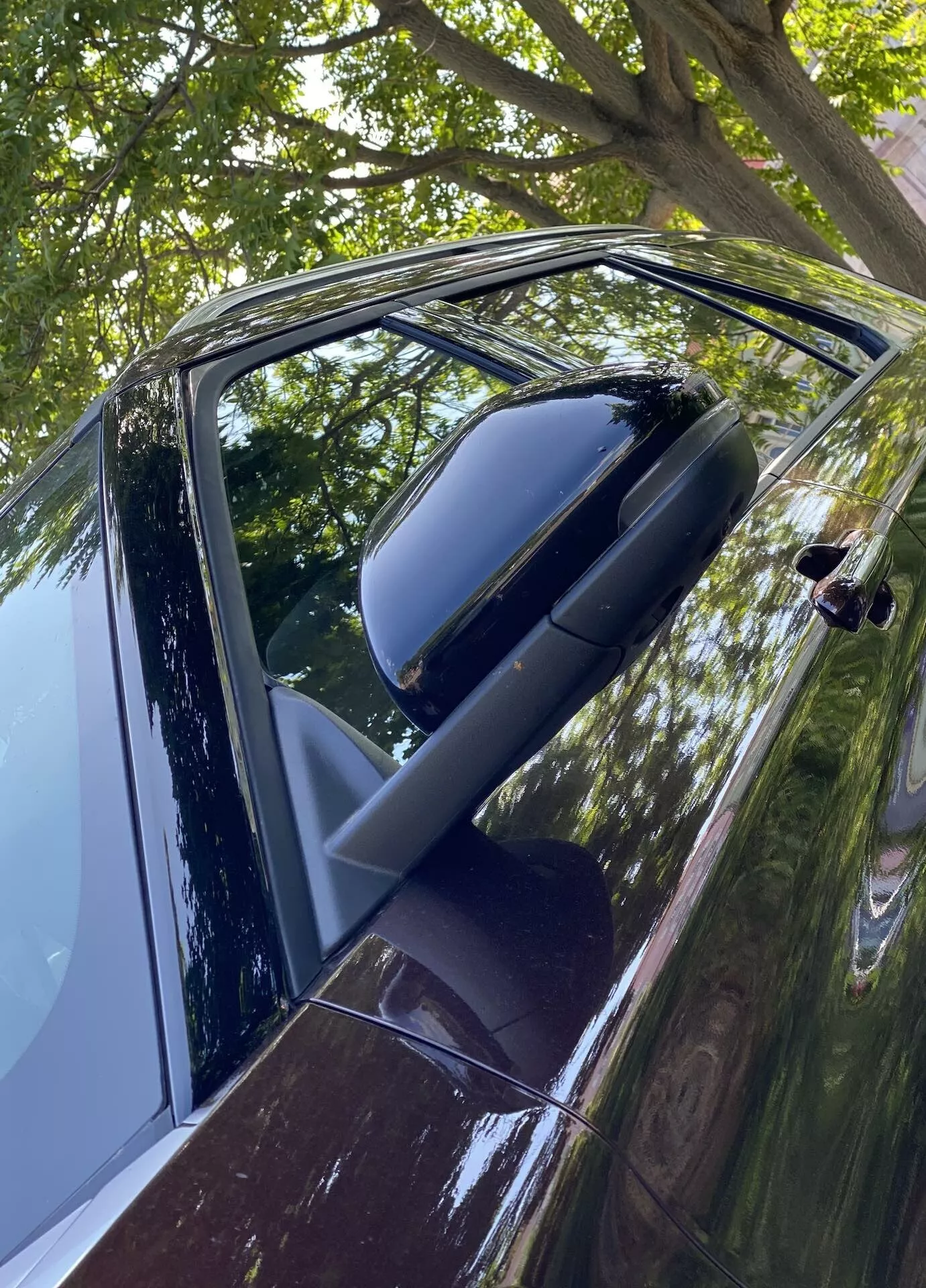 Närbild, vinklad vy av förarsidans dörr och sidobackspegel på en Ford Explorer 2019. Bilden, tagen något ovanför dörrhandtagsnivå, visar upp en glansig mörk exteriör, möjligen svart eller djupblå, med reflektioner som indikerar en ren, välskött kaross. Den ihopvikta spegeln, färgkoordinerad med karossen, antyder en högre modell. Delvis nedvevad ruta och modern, möjligen beröringssensor utrustad dörrhandtag, antyder nyckellös in- och utgångsteknik. Reflektioner av löv och grenar tyder på utomhusparkering.