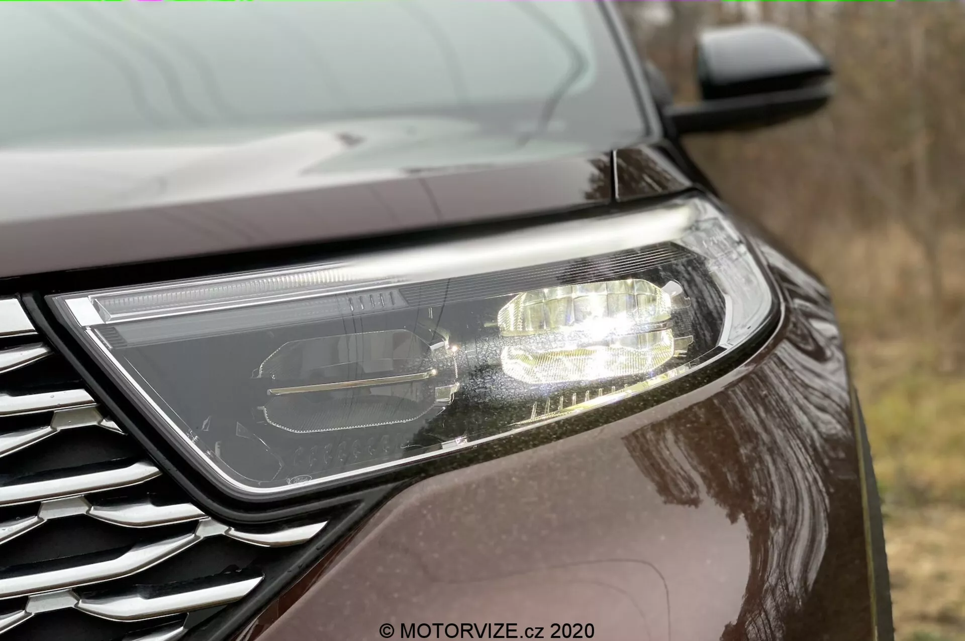 Närbild från sidan av fronten på en Ford Explorer 2019. Framträdande modern strålkastare som antyder LED eller HID-teknik med aktiva dagsljuslampor. Grillen är delvis synlig med en metallisk emblem, placerad i en låg vinkel. Bilen har en mörk, reflekterande lackfinish som antyder en metallisk eller pärlemorkvalitet, i en utomhusmiljö.