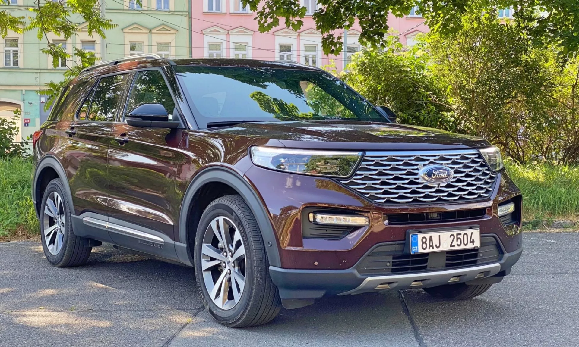 Trekvart frontvy av en brun Ford Explorer SUV från 2019, som visar dess sidoprofil och frontparti. Bilen är parkerad vid en stadsgata med byggnader och träd i bakgrunden. Här syns flera nyckelfunktioner, inklusive en framträdande frontgrill med horisontella slitsar och centralt placerad logotyp, moderna LED-dagljus, dimljus integrerade i det nedre stötfångarområdet, stora mångekrade aluminiumfälgar, sidospeglar med integrerade blinkers, tonade fönster, takräcken, konturerade karosslinjer och en hög markfrigång.