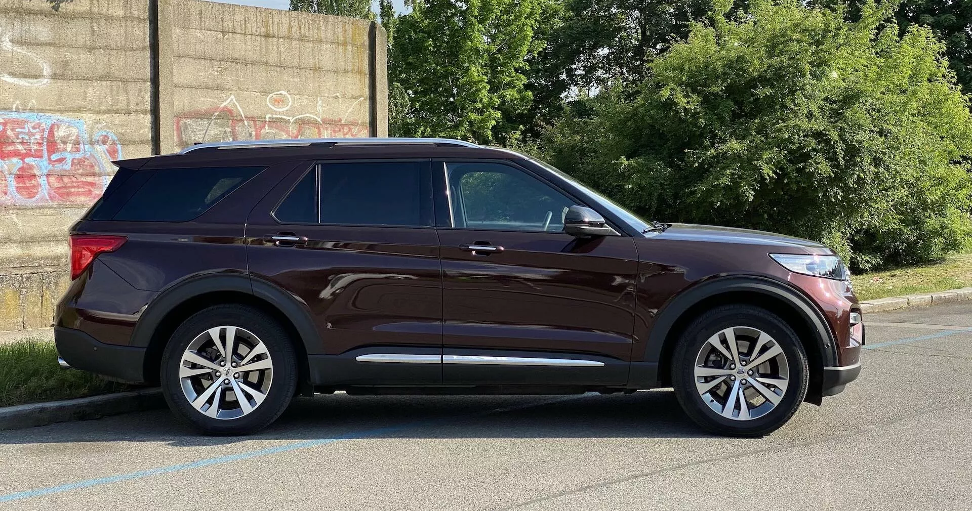 Moderna SUV:n Ford Explorer 2019 i sidovy, parkerad på ett asfalterat område bredvid en trottoarkant med en graffiti-täckt vägg i bakgrunden som antyder en urban miljö. Egenskaper inkluderar eleganta strålkastare, framträdande karaktärslinje, sidospeglar med indikatorer, femekrade aluminiumhjul, kromdetaljer runt fönstren, takräcken, tonade bakrutor, kroppsfärgade dörrhandtag och gott om markfrigång. Dagsljusfotografering visar hela fordonets profil.