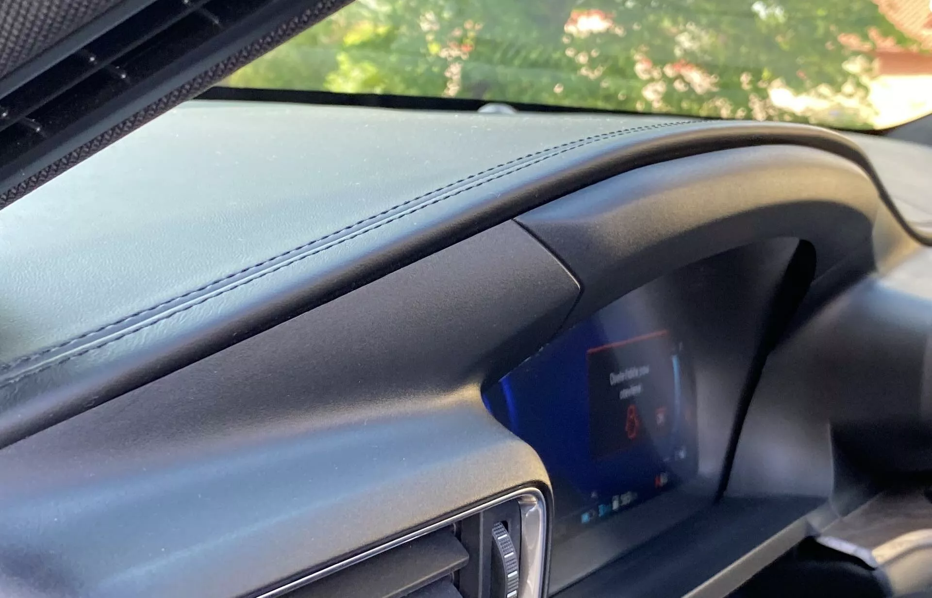 Interiörvy av en Ford Explorer 2019 från passagerarens perspektiv som visar en modern instrumentbräda med läderliknande textur och framträdande horisontella sömmar samt en digital instrumentpanel med blå skärm och röda varningsindikatorer. Den mörkgrå och svarta färgschemat samt delvis vy på dörrens ram indikerar ett premiumfordon i en väl upplyst miljö.