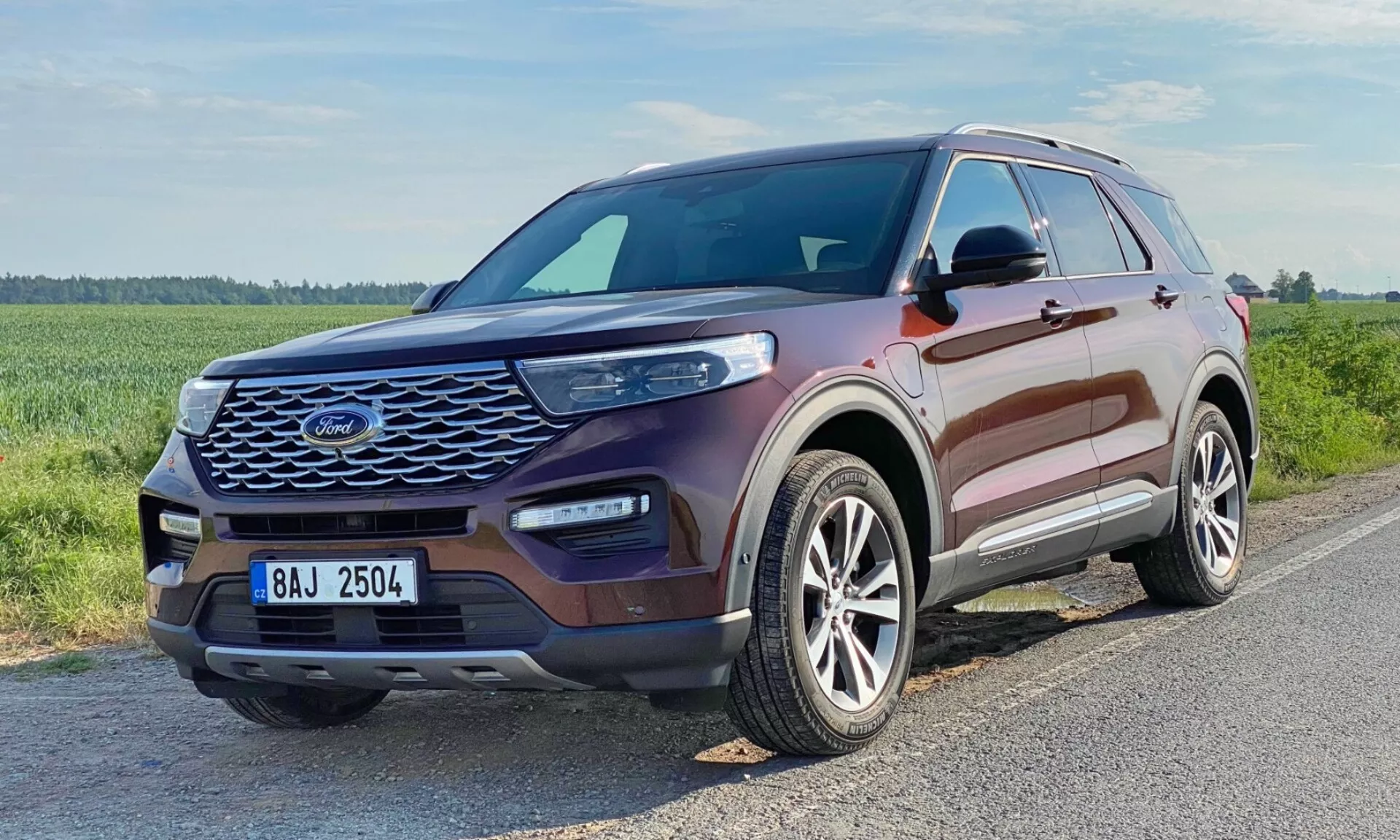 Tre-kvartiers vy framifrån av en Ford Explorer SUV 2019. Bilden visar fordonets främre grill med ett nätliknande mönster, eleganta långsträckta strålkastare med synliga LED-dagljuslampor, en skulpterad motorhuv med upphöjda konturer, lättmetallfälgar med en mångekerad design och färgmatchade sidospeglar med integrerade blinkers. Fönstren har krom- eller silverfärgad trim. Fordonet är metalliskt kastanjebrunt och placerat på en asfalterad väg i en gräsbevuxen, möjligen landsbygdsmiljö.