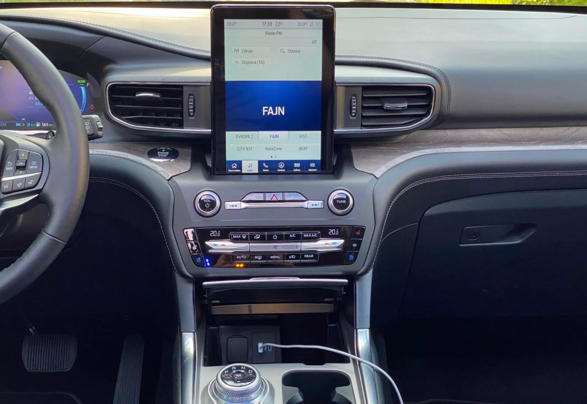 Interiören av en Ford Explorer 2019, som visar den moderna, centrala instrumentpanelen från en fram- eller baksätesperspektiv. Instrumentpanelen har en upprätt infotainment pekskärm med klimatkontroller nedanför, inklusive inställningar för dubbla klimatkontroller, en CD-kortplats och medieinpålningsalternativ. Den centrala konsolen inkluderar en växelspak med metallaccent, kontrollknappar, en roterande ratt och mugghållare. Materialen antyder en blandning av träkorn, metalliska ytbehandlingar och svart plast, och framhäver fordonets teknik och kvalitativa interiördesign.