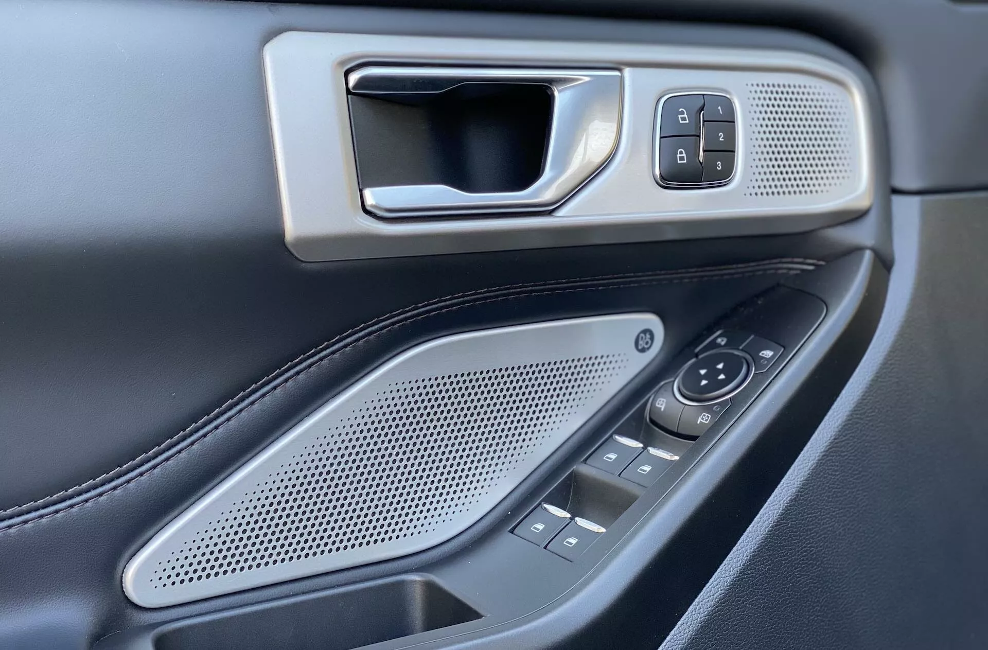 Bilden fångar förarsidans interiöra dörrpanel i en Ford Explorer 2019. Fokuspunkten från en något förhöjd och vinklad perspektiv visar ett metalliskt utseende dörrhandtag inbäddat i en ram, bredvid lås/öppningsknappen inbyggd i en fint texturerad panel. Nedanför finns ett ovalt högtalargaller med runda hål och flera kontrollknappar och strömbrytare för sidospeglar och fönster, vilket antyder eldrivna fönster för flera dörrar. Den mörka färgpaletten med kontrasterande metalliska element och synliga sömmar på en troligtvis läderklädd armstöd förbättrar lyxkänslan. Det grunda skärpedjupet framhäver kontrollelementen, vilket betonar design och placering.