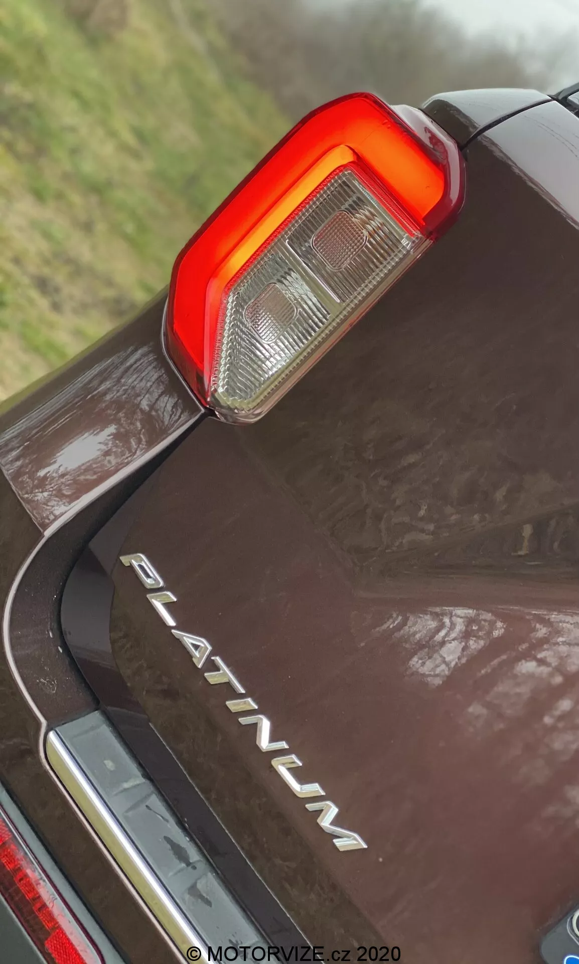 Närbild i låg vinkel av bakre delen på en 2019 Ford Explorer PLATINUM, fokuserad på den vertikala LED-baklyktan med klara och röda sektioner för back- och bromsljus, krom 'PLATINUM'-märke, reflekterande mörk färg med vattendroppar, och kromdetalj under bakre stötfångaren.