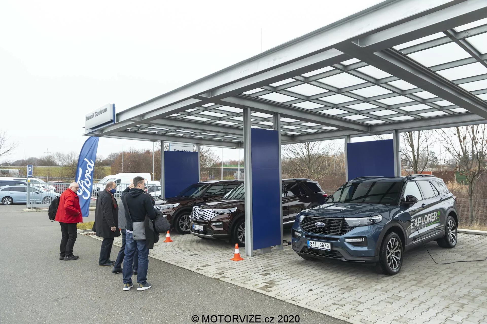 Bilden visar en utomhusutställning eller bilförsäljning under en metallram med ett semitransparent tak i mjukt, övermulet dagsljus. Tre SUV:ar, inklusive en modern Ford Explorer 2019 i förgrunden, visas upp med synliga funktioner som frontgaller, strålkastare och sidospeglar. Folk och affischer pekar på ett officiellt promotionevenemang, och Ford-logotypen är synlig i bakgrunden.