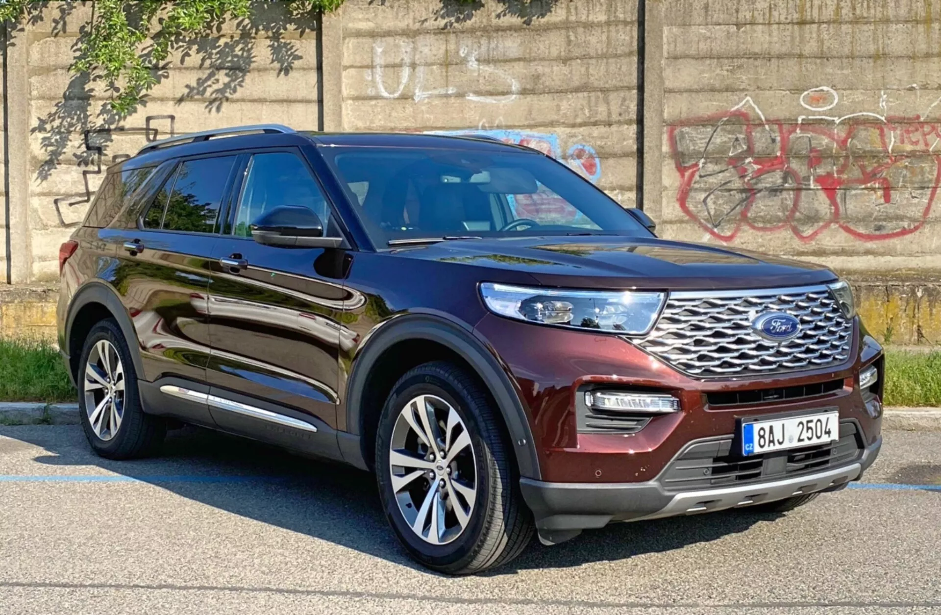 Bild på en 2019 Ford Explorer SUV tagen från en trekvarts frontal vinkel på förarsidan, visar en blank metallisk brun eller vinröd färg. Bilden framhäver den främre grillen med Ford-logotypen, eleganta LED- eller HID-strålkastare, främre stötfångare och förarsidans profil inklusive dörrar, stänkskärm och flerspakiga aluminiumhjul. Fordonet är parkerat i ett stadsområde med graffiti på en betongvägg i bakgrunden, vilket framhäver dess robusta och moderna design, med möjliga avancerade förarassistanssystem såsom adaptiv farthållare och filhållningsassistans.