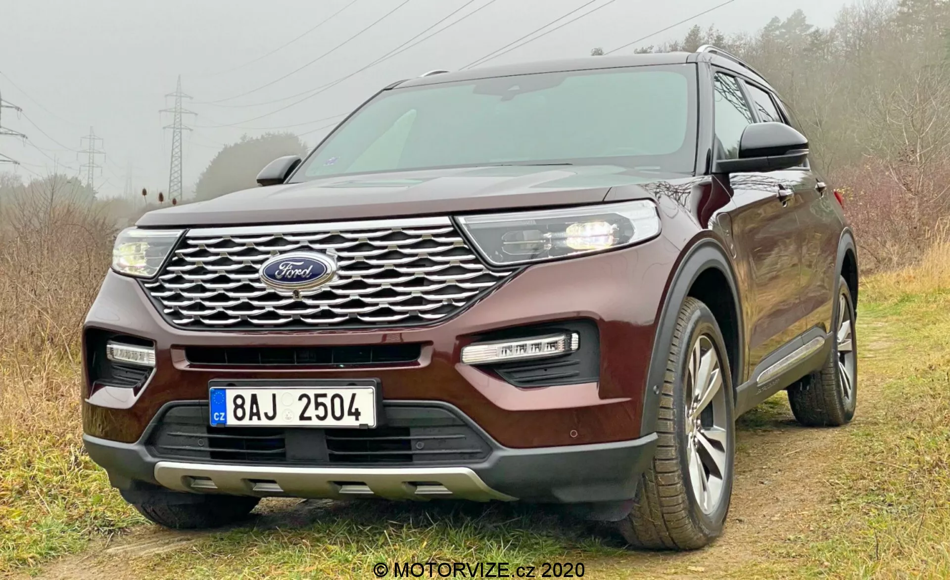 2019 Ford Explorer bil fotograferad från en låg front-trekvarts vinkel som visar grillen med Ford-logotypen, strålkastare, dimljus, motorhuv, sidorutor och en del av bakdelen. Grillens intrikata design, eleganta LED-strålkastare, tvåfärgad stötfångare och nyckellöst inträdesystem framhäver dess moderna och teknologiskt avancerade funktioner. Placerad något off-road med höger hjul på gräs och vänster hjul på jord, vilket framhäver lätt off-road kapacitet. Flerspakiga legeringshjul synliga, vilket tyder på året-runt däck.