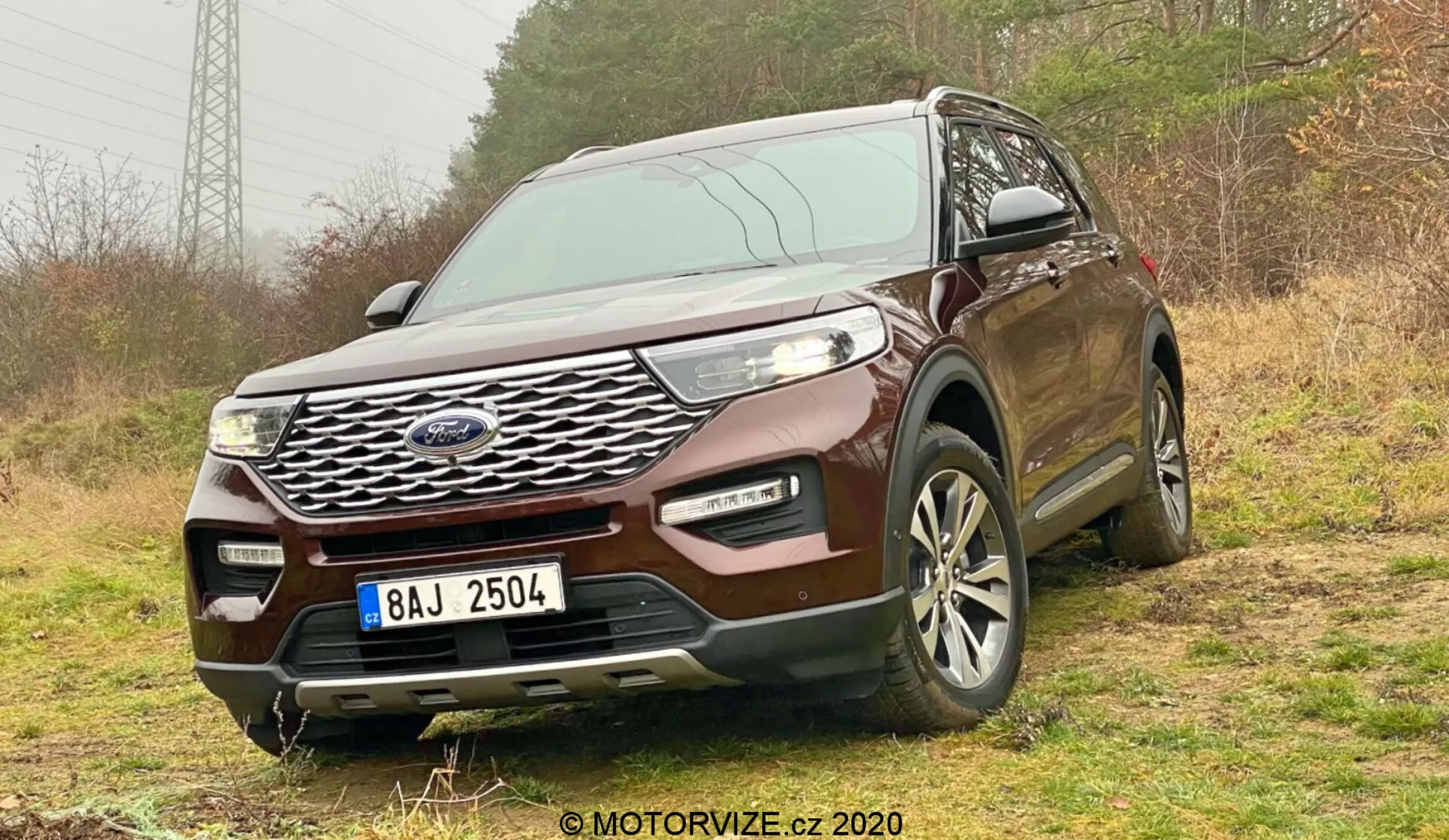 Frontal trekvartsvy av en 2019 Ford Explorer SUV tagen från en låg vinkel, som visar dess framträdande kromgrill, eleganta LED-strålkastare och flerspakiga aluminiumfälgar. SUV:n är placerad på en oförseglad yta med vegetation, vilket antyder dess terrängkapacitet. Den främre stötfångaren inkluderar dimljus och luftintagsgaller, vilket tyder på förbättrad funktionalitet. Element som sensorer eller kameror kan indikera avancerade säkerhetsfunktioner. Registreringsskylten är censurerad och ett vattenmärke lyder '© MOTORVIZE.cz 2020'. Vinkeln betonar dess robusta hållning och tekniska sofistikering.