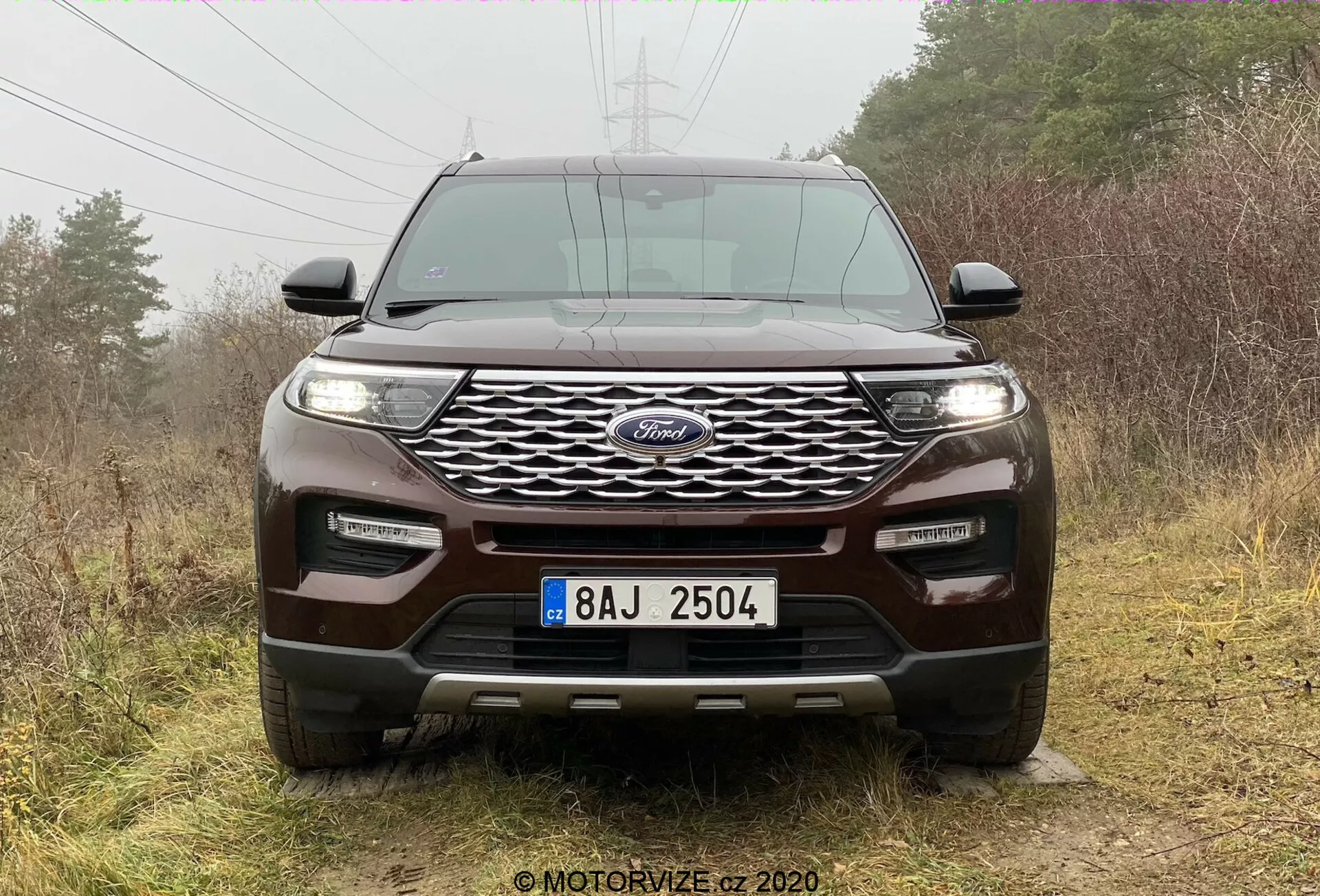Frontal vy av en 2019 Ford Explorer SUV som visar ett framträdande blått ovalt emblem centrerat på det dynamiska vågiga grillmönstret, flankerat av eleganta horisontellt orienterade strålkastare sannolikt med LED- eller HID-ljus. Integrerade dimljus och parkeringssensorer förbättrar framstötsbulletens teknologiska och säkerhetsfunktioner. Den skulpterade motorhuven med upphöjda åsar betonar den sportiga designen, medan den stora markfrigången, skyddsplåten och bogserhakens indikerar terrängförmåga. SUV:en är placerad mot en utomhus, naturlig bakgrund med diffust molnljus som framhäver dess design.