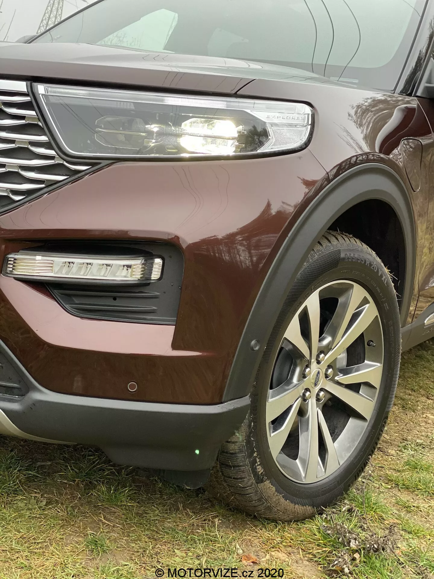 Närbild i vinkel av en 2019 Ford Explorer SUV med fokus på det vänstra främre kvartalet, som lyfter fram dess stora lättmetallhjul, grill med kromaccenter och delvis synlig LED-strålkastare med dagljus, metallic vinröd lack, dimljus i det inbäddade stötfångarområdet med annan textur och eventuellt luftintag eller dekorativ trim. Logotypen är i mitten av de flerkantiga hjulen. Den nedre stötfångardelen antyder närvaron av en parkeringssensor eller ett adaptivt farthållarsystem. Bilden är tagen ur en låg vinkel för att framhäva fordonets storlek och robusthet, med en vattensignatur 'MOTORVIZE.cz 2020' synlig i nedre högra hörnet.