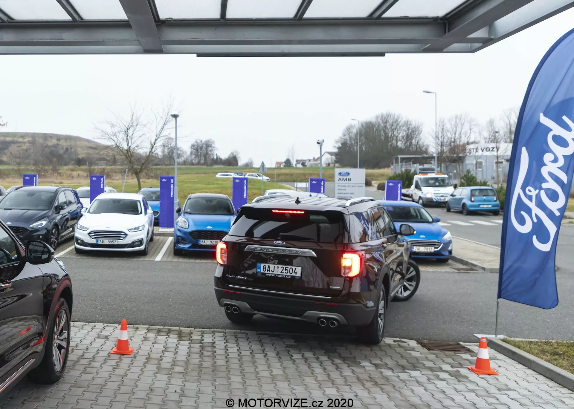 En svart 2019 Ford Explorer SUV, placerad centralt, backar antingen in i eller lämnar en laddplats för elbilar vid en laddstation. Bakljusen är på, möjligen indikerande backning eller inbromsning, med en synlig registreringsskylt, dubbla sportavgassystem, bakre fönstertorkare och högt monterad bakre bromsljus. Omgivningen visar flera elbilar vid blå laddstolpar och orange trafikkoner nära SUV:en. En flagga med Ford-logotypen antyder varumärkesnärvaro. Scenen är fångad en molnig dag med diffust naturligt ljus, vilket framhäver fordonskonstruktion och hållbar transportinfrastruktur.