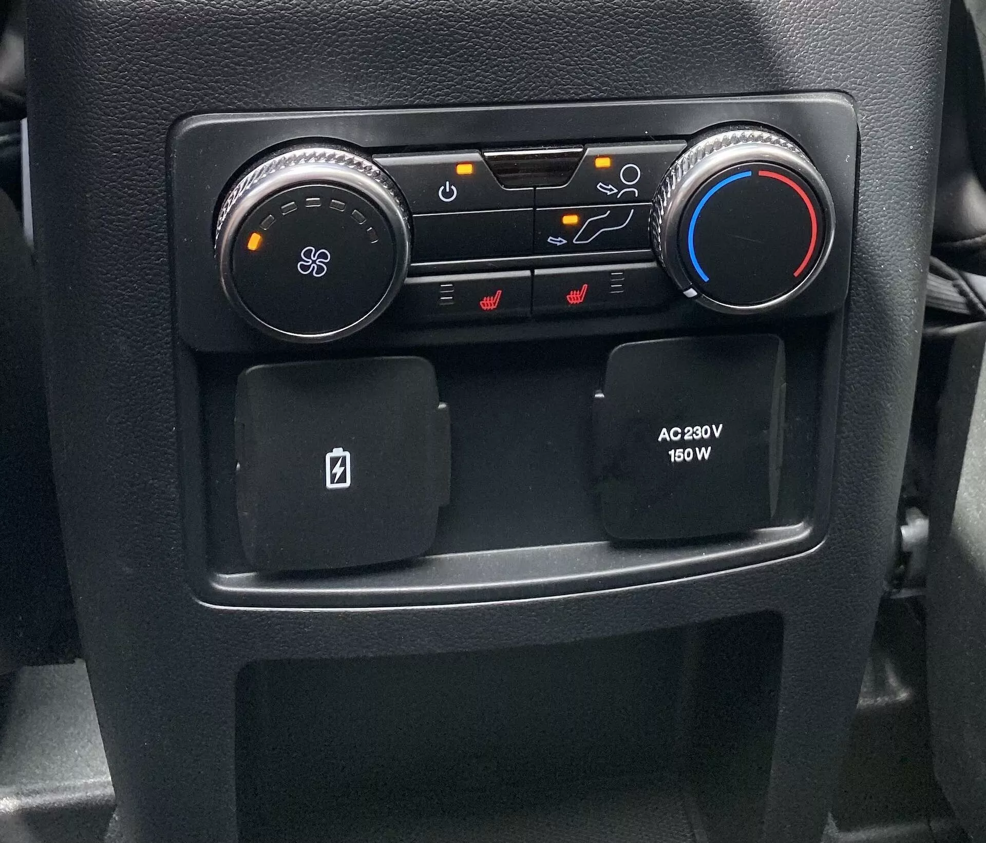 Närbild av en sektion av mittkonsolen i interiören på en Ford Explorer 2019, som visar HVAC- och komfortkontroller. Till vänster finns en rund svart ratt med en fläktikon som justerar blåsfunktionen; bredvid den finns knappar för luftflödesriktning, luftcirkulation och avfrostningsfunktioner för vindrutorna. En blå och röd ratt till höger reglerar temperaturen. Nedanför finns två halkskyddade förvaringsfack, ett utformat för nyckelkort, och ett eluttag på passagerarsidan som erbjuder 230V och 150W för elektroniska enheter.