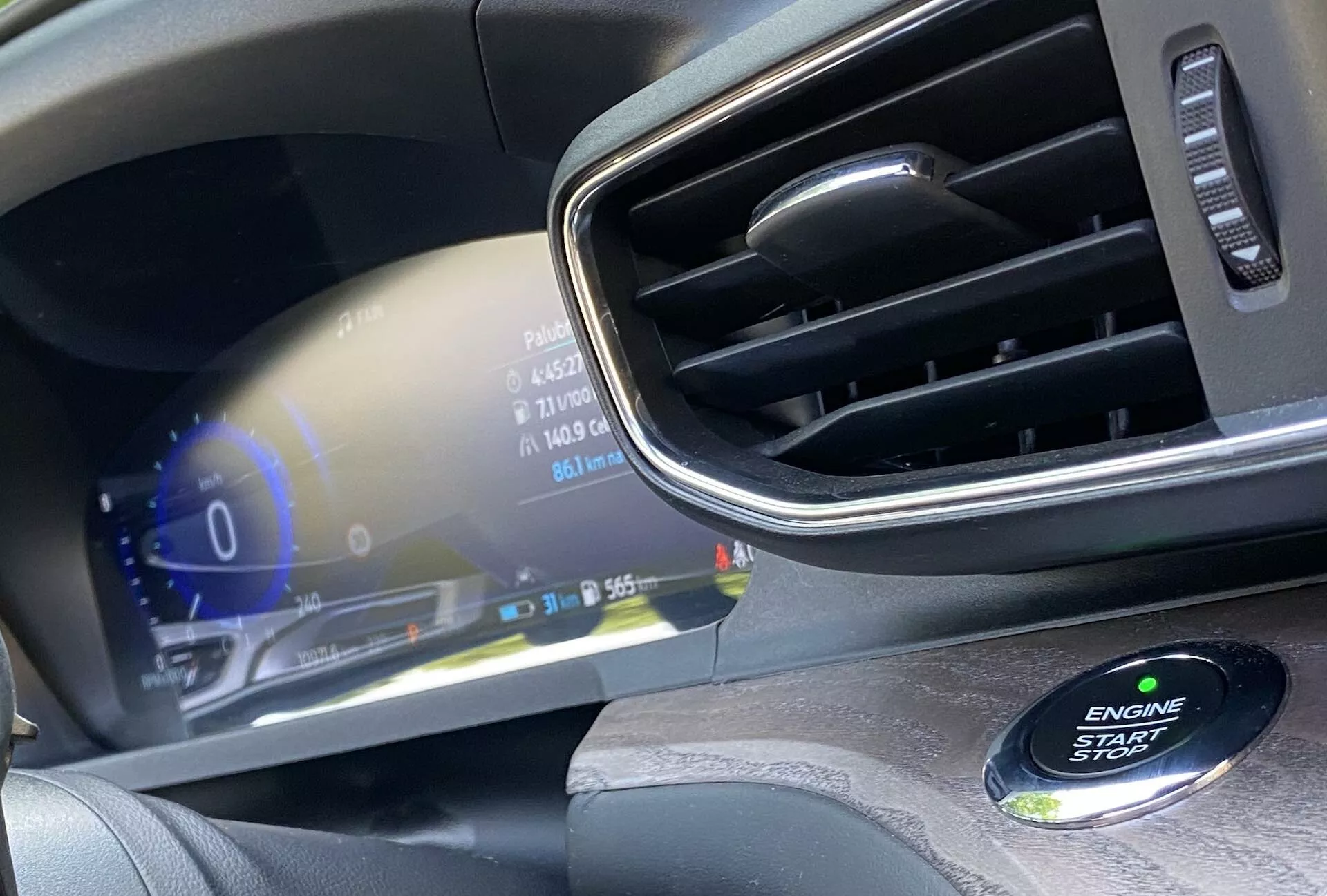 Närbild av interiören i en modern bil, med tonvikt på förarsidans instrumentbräda. Bilden betonar den digitala instrumentpanelen som visar olika fordonsstatusar, såsom hastighetsmätaren på 0 km/h, bränslemätaren och utomhustemperaturen som visar 31 grader Celsius. Den högupplösta skärmen har livfulla färger och en lyxig design. Till höger syns kanten av en luftventil med kromdetaljer, och i det nedre högra hörnet syns, något ur fokus, en ENGINE START STOP-knapp som antyder ett nyckellöst startsystem.