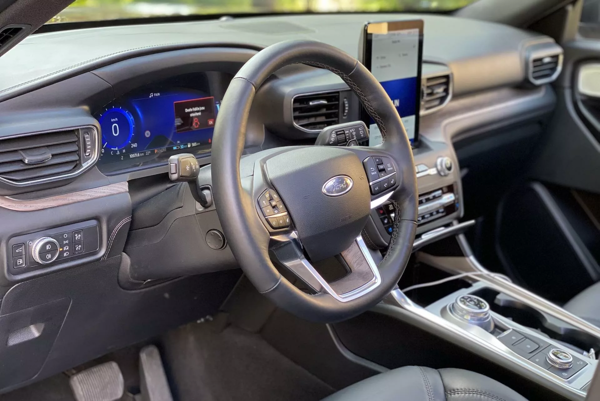 Interiören av en 2019 Ford Explorer med fokus på förarområdet från främre passagerarens perspektiv. Ratten, med multifunktionskontroller för ljud, farthållare och handsfree-kommunikation, är centrerad. Bakom den finns en digital instrumentpanel som visar anpassningsbara mätare och realtidsfordonsdata. Till höger ser vi en vertikalt orienterad pekskärm som visar ett infotainmentsystem med satellitnavigering och smartphone-integration. Nedan finns flerzons klimatanläggningskontroller och en roterande växelväljare, vilket indikerar en modern design. Kabinen har en ren och modern instrumentpanel tillverkad av premium-material och utstrålar en minimalistisk estetik.