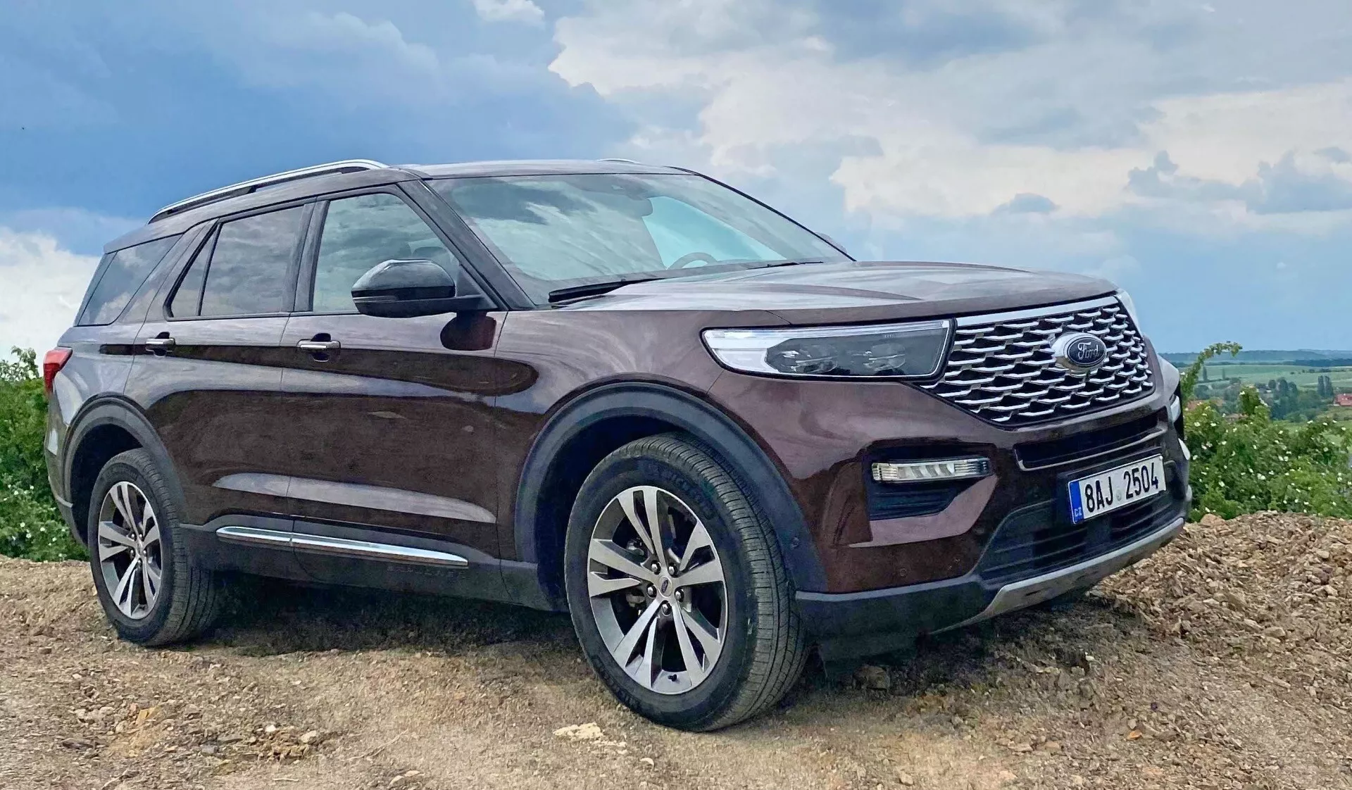 Bild på en Ford Explorer SUV från 2019 parkerad på en lutande, möjligen oasfalterad yta med mörk nyans och smutsstänkta sidor, framträdande grill med nätmönster, LED-strålkastare, dimljus, legeringshjul med flerspakigt mönster, sidospeglar med integrerade blinkers, takräcken, sidosteg, lätt tonade fönster och en antenn på bakre taket, som betonar dess robusta kapacitet och lyxiga egenskaper i en lantlig eller off-road-miljö.