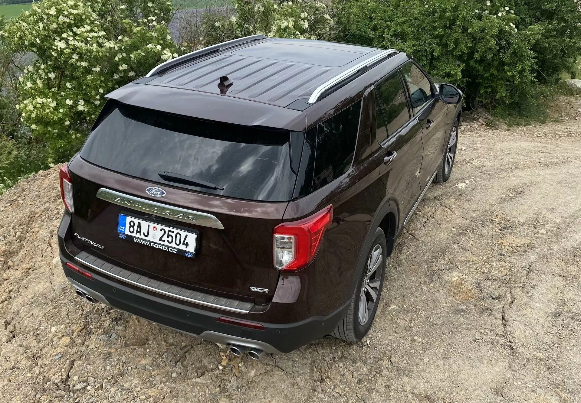 Upphöjd bakifrån-snett-vy av en mörkfärgad 2019 Ford Explorer, som framhäver fordonets bakre ände och sidoprofil. Synliga funktioner inkluderar Ford-logotypen, modellnamnet 'Explorer', unika bakljus, bakre vindrutetorkare, hajfenaantenn, dubbelt avgasrörssystem, backkamera, lättmetallfälgar, tonade rutor och sidobackspeglar med integrerade blinkers. SUV är något förskjuten åt höger på ojämn terräng, vilket antyder lätt terrängförmåga.