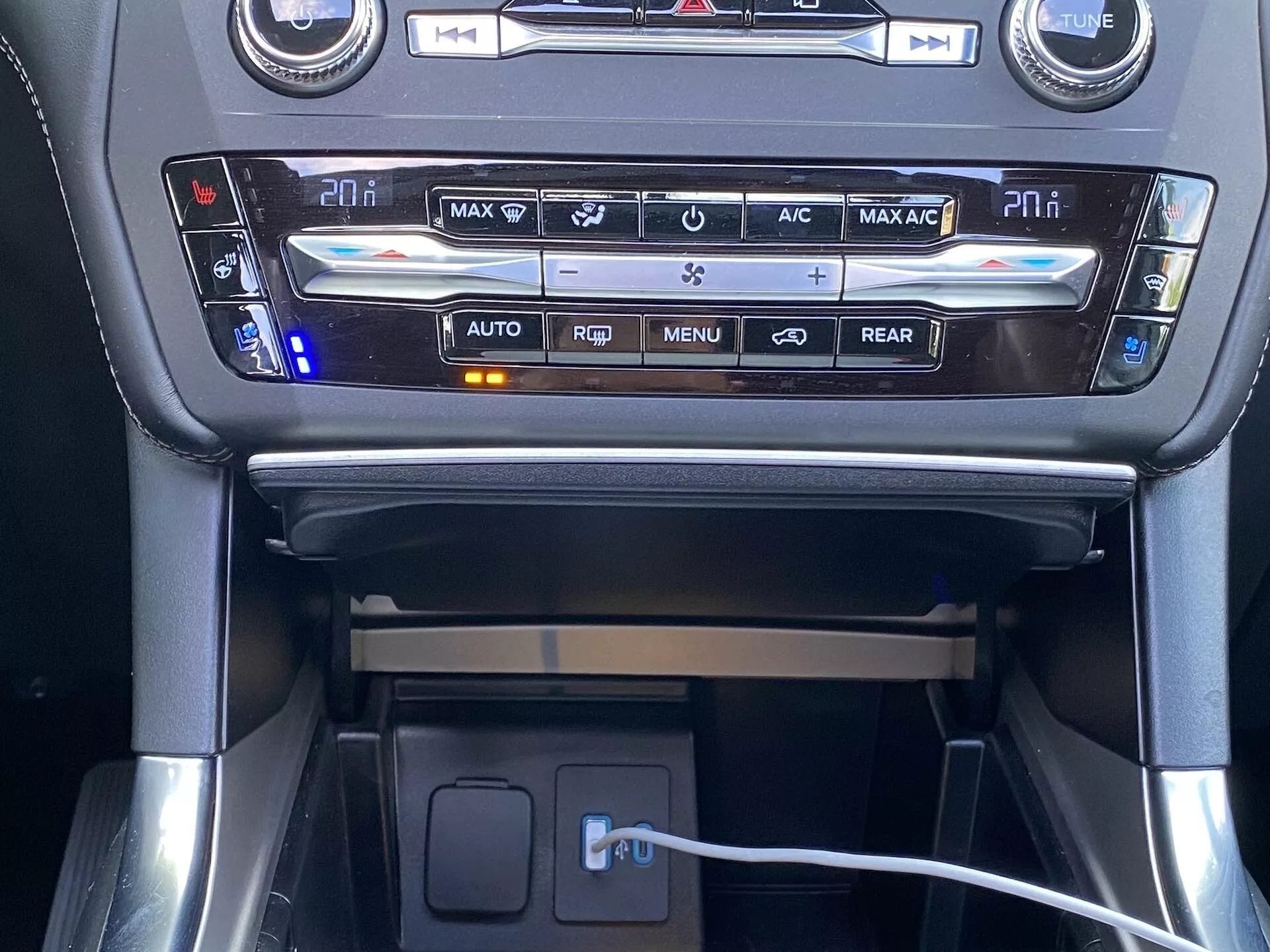 Främre interiöruppfattning av mittkonsolen i en Ford Explorer 2019, fokuserad på klimatanläggningen och förvaringsutrymmet. Den välbelysta klimatpanelen innehåller digitala temperaturvisare för förare och passagerare, omgiven av knappar för olika funktioner inklusive avfrostning, luftcirkulation och automatisk klimatkontroll. Under detta panel finns en liten gummerad hylla med en synlig laddningskabel som indikerar anslutnings- eller laddningsfunktioner.