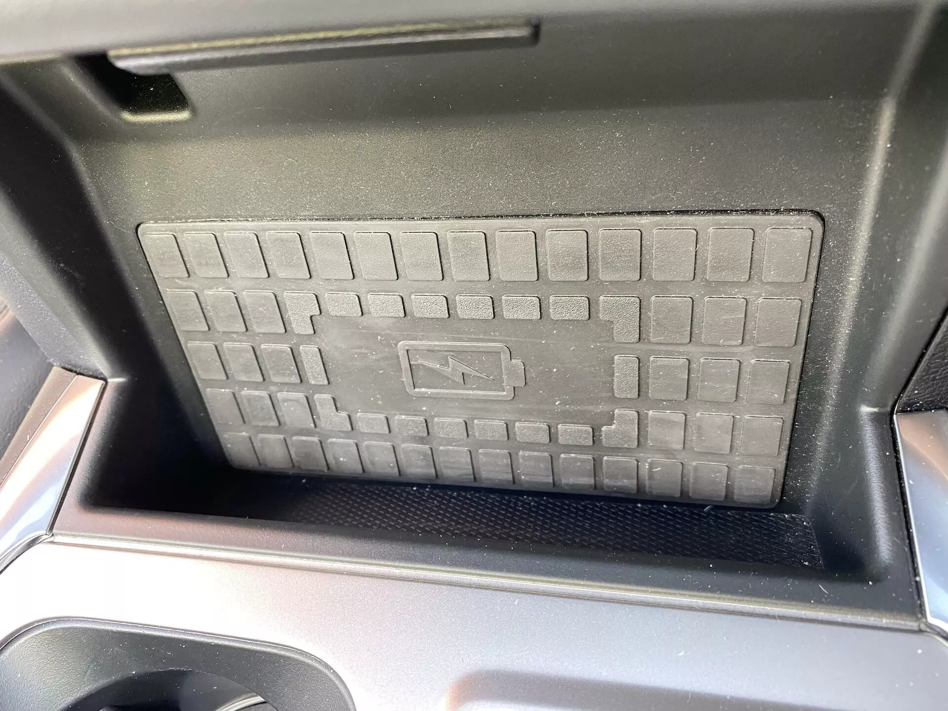 Närbild av mittkonsolfacket i Ford Explorer 2019, med fokus på den trådlösa laddplattan med upphöjd rutnätsstruktur och halkfri yta. De glänsande svarta plastomgivningarna är synliga, vilket ger en touch av modern bildesign. Solljuset antyder att fotot är taget på dagen från framsätet, med laddplattans symbol som anger avancerad trådlös laddningsteknik.