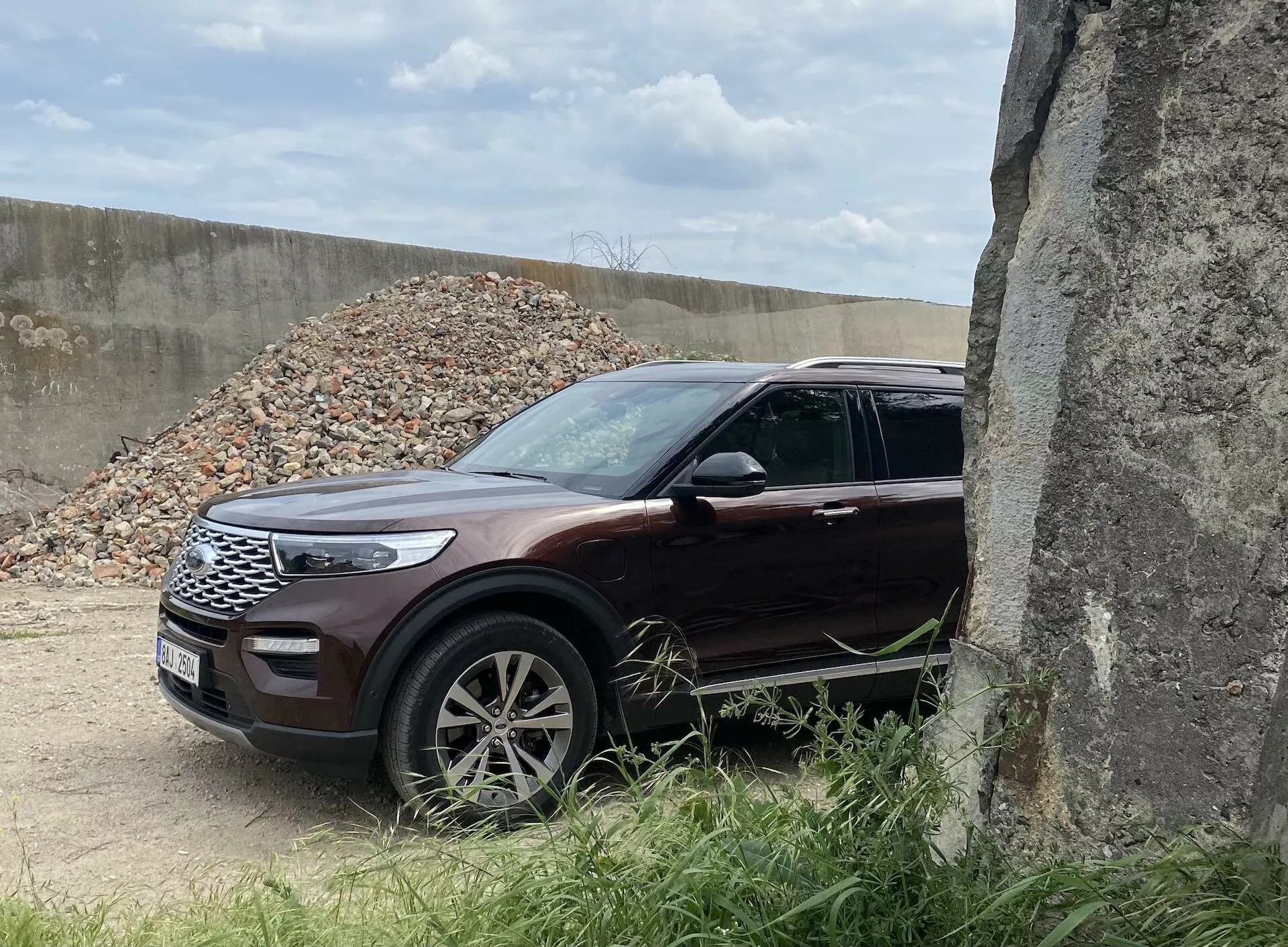 Bilden visar en Ford Explorer SUV från 2019 ur en trekvarts främre vy, som framhäver den vänstra främre grillen, eleganta LED-strålkastare, motorhuv, framhjul och skärm. Fordonet, i en glansig djup mörkröd eller vinröd färg, står i en utomhusmiljö med grus i bakgrunden, vilket antyder en byggarbetsplats. Flerekrade lättmetallfälgar, integrerade blinkers i speglarna och kromdetaljer förstärker det premiumutseendet. Delvis skymd av en betongstruktur till höger, uppvisar denna SUV moderna design- och säkerhetselement, vilket gör den lämplig för kategorier som SUV, modern bildesign, fordons teknologi och terrängfordon.