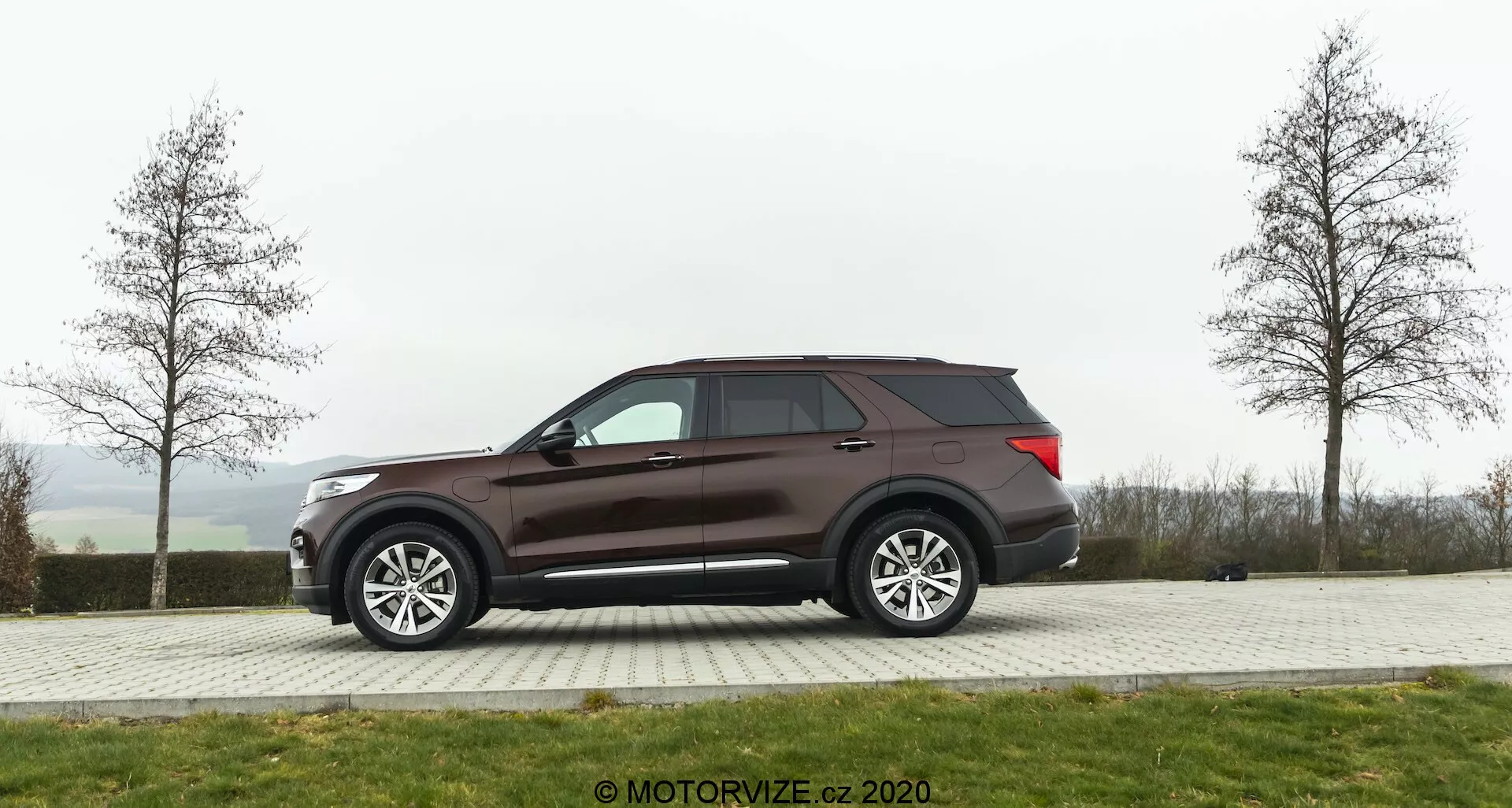 Sidoprofil av en Ford Explorer SUV från 2019 parkerad på ett asfalterat område med gräs i bakgrunden på en mulen dag. Fordonet har stora fler-ekrade lättmetallhjul, tonade rutor och en mörkbrun eller vinröd lack. Designdetaljer inkluderar sidospeglar med troligen elektrisk justering och integrerade blinkers, aerodynamisk taklinje och en synlig bakantenn.