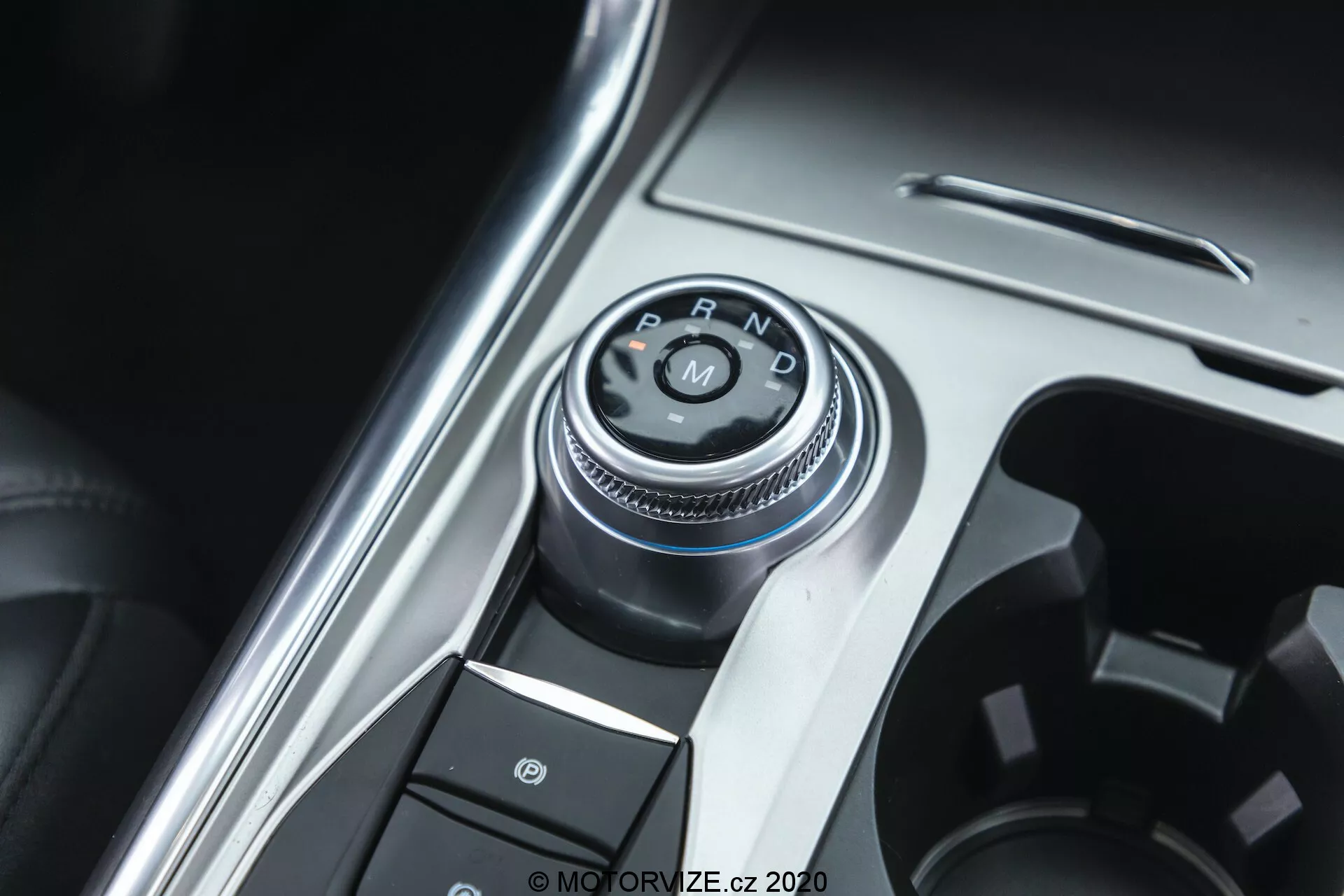 Närbild av växelväljarknappen och en del av mittkonsolen i en Ford Explorer från 2019. Huvudpunkten är den roterande växelväljaren med 'P, R, N, D'-etiketter som indikerar automatiskt växellåssystem. Fotot är taget från en något sned, uppifrån-och-ner-vinkel. Runt växelväljaren finns knappar, inklusive en med en parkeringsbromssymbol, vilket tyder på ett elektroniskt parkeringsbromssystem. Konsolen kombinerar metalliska och plastiga ytor som betonar fordonets premiuminteriör. Den välbelysta bilden framhäver de metalliska elementen med ett grunt skärpedjup fokuserat på växelväljaren.