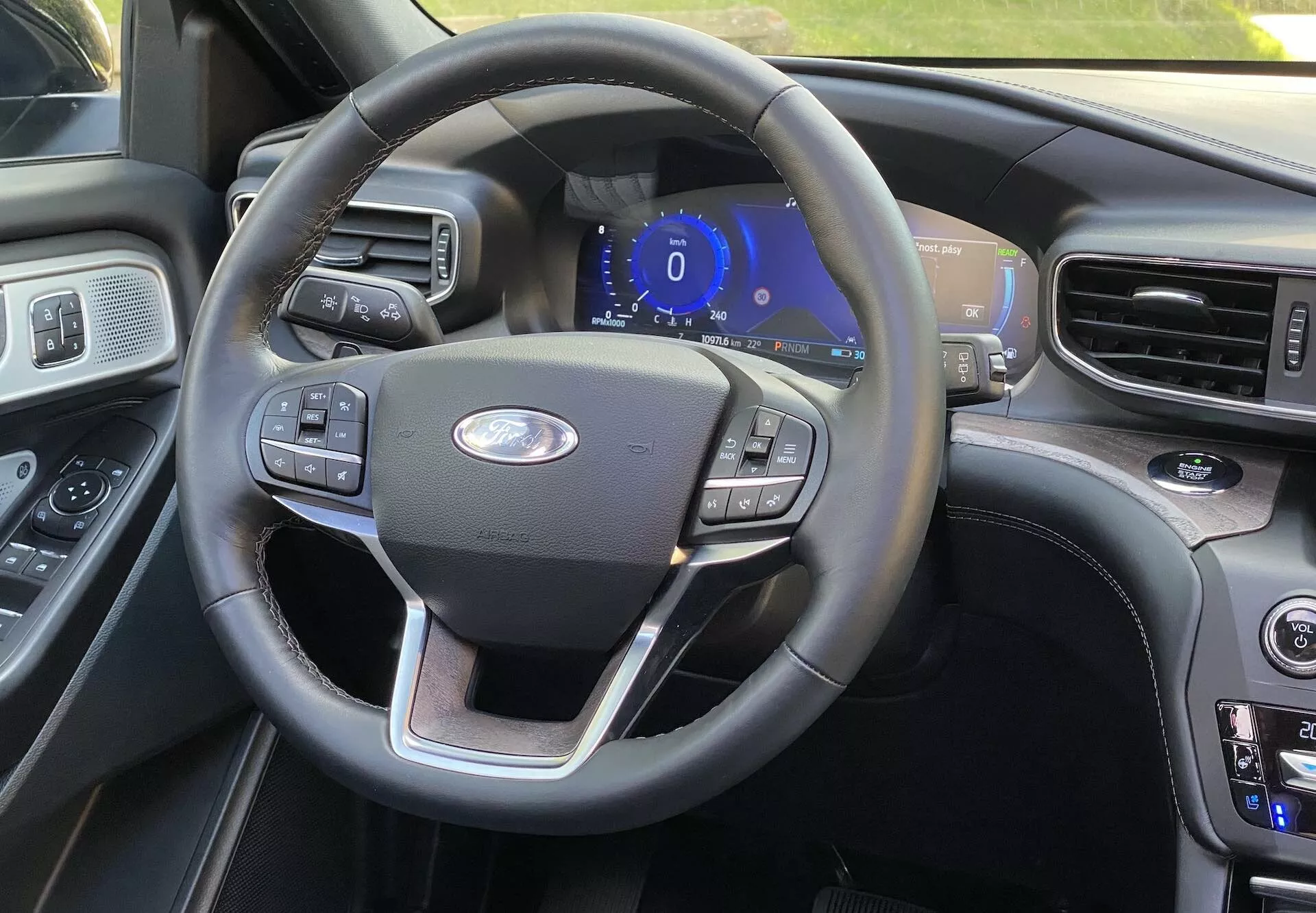 Interiörvy av en Ford Explorer 2019 från förarsätet, som visar en treekrad ratt med multifunktionella knappar för farthållare, ljudinställningar och telefonanslutning. Det centralt placerade Ford-emblemet och en blåbelyst digital instrumentpanel antyder att fordonet är i tillbehörs- eller startläge. Till höger syns en del av mittkonsolen med klimatanläggningsknappar och temperaturinställningar, vilket tyder på dubbel klimatkontroll. Interiören har en blandning av läder och träkornfinish för en premiumkänsla, med en annan större skärm lätt synlig i kanten.
