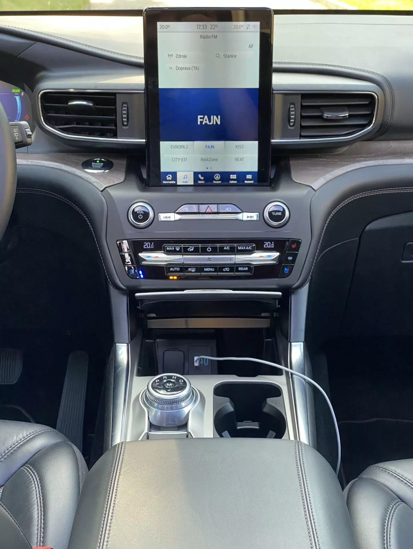 Interiörvy av en 2019 Ford Explorer från perspektivet av en passagerare i baksätet, med fokus på det främre kabinområdet med ett avancerat infotainmentsystem på mittkonsolen, tvåzons klimatkontroll, roterande växelväljare och lyxiga detaljer i läder och krom.