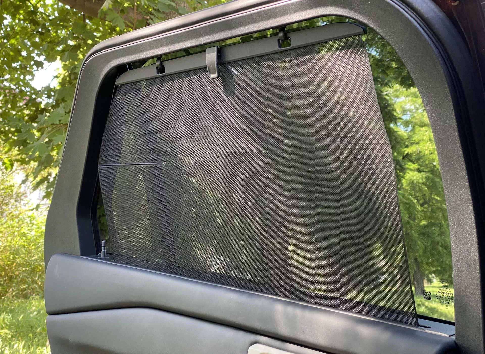 Närbild av interiören på Ford Explorer 2019:s bakre sidodörr och fönster. Bilden fångar ett delvis nedrullat fönster med en nät-solskydd installerat, som täcker hela glasytan och sticker ut något över den övre kanten. Den övre delen av bilens dörrpanel med slät ytbehandling är också synlig. Fotot, taget i horisontell orientering, visar ljus som filtrerar genom solskyddet, vilket antyder en park-liknande utomhusmiljö. Fokus på passagerarkomfort med solskyddstillbehör.