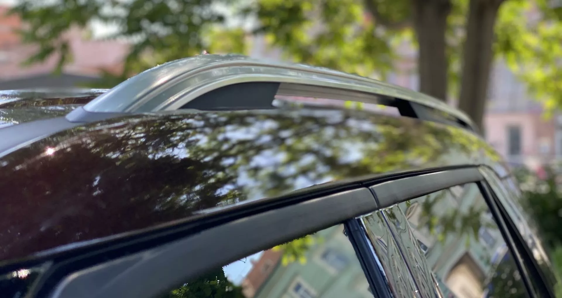 Närbild av taket på en Ford Explorer 2019 från låg vinkel, som visar det panoramiska soltaket i ventilerad position, metalltrim runt fönstren och takräcken mot en suddig bakgrund av träd och arkitektur.