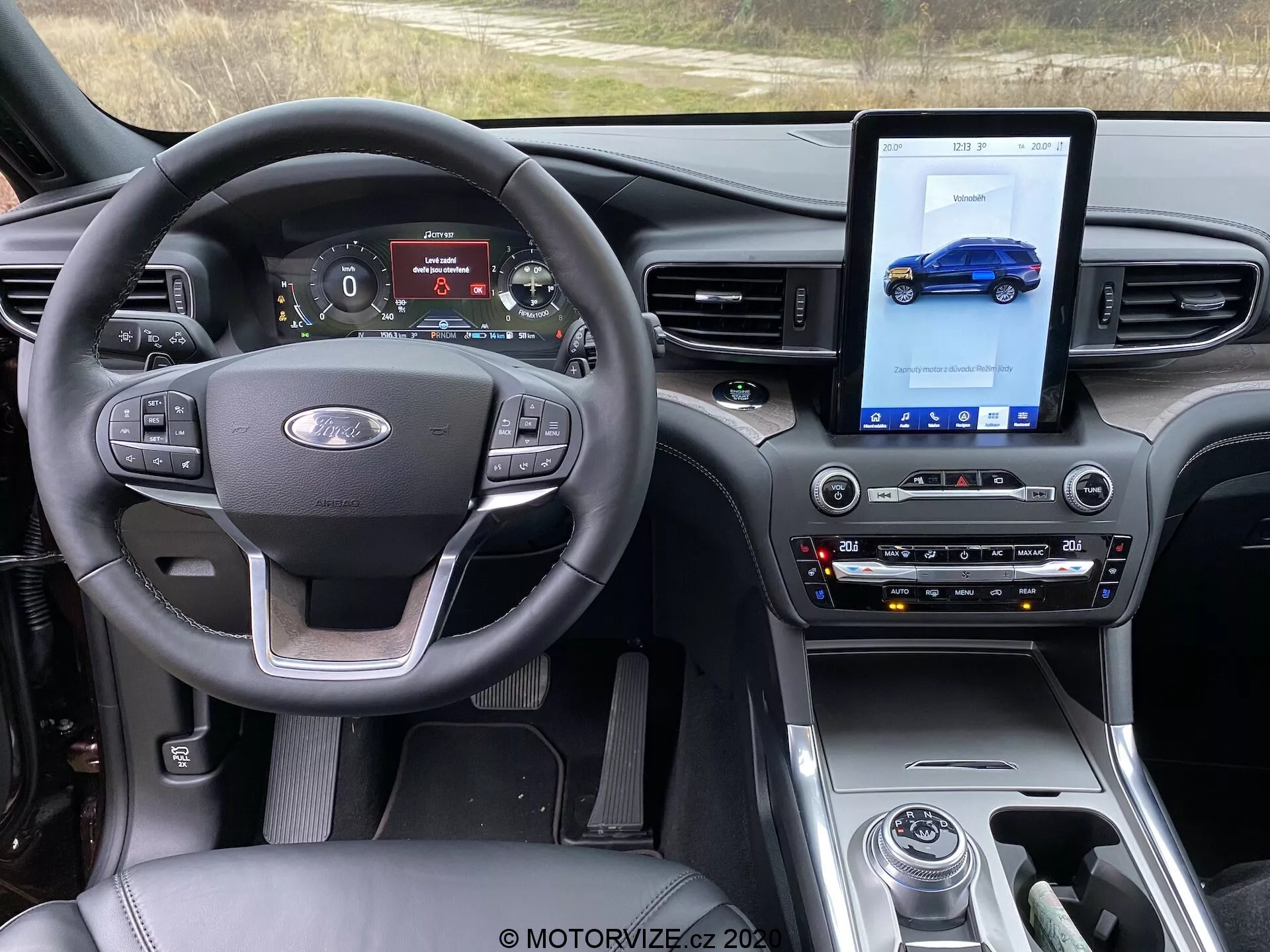 Interiör av en Ford Explorer 2019 med fokus på framsätet ur förarens perspektiv. Bilden visar en ratt med kontrollknappar för volym, röstkommandon och farthållare, tillsammans med en Ford-logga i mitten. Den digitala instrumentpanelen visar hastighetsmätare, varvräknare, körläge och bränslestatus. Mittkonsolen har en stor vertikal pekskärm för infotainmentsystemet, som visar en illustration av ett blått fordon. Tradionella kontrollpaneler finns för klimatanläggning och ljudsystem, och en växelväljare syns också. Instrumentbrädan har en modern design med blanka ytor, matta finishar och inslag av borstad metall, vilket förstärker den moderna, premiumkänslan. Tekniska funktioner inkluderar multifunktionell ratt, digital instrumentpanel, pekskärms infotainmentsystem, automatisk klimatanläggning, körlägesväljare och sätesvärme reglering.