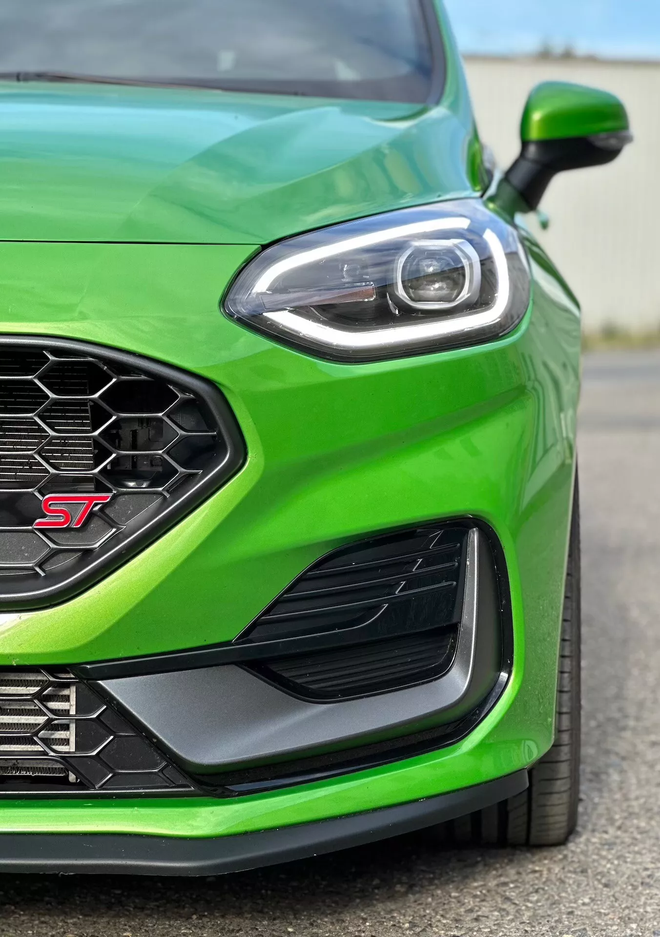 Närbild av den prestandainriktade främre vänstra kvarten av en Ford Fiesta ST 2022 i livlig grönt, belyser den sportjusterade trimmen, bikakeformad grill för optimal luftflöde, avancerad LED-strålkastardesign, aerodynamisk sidoventilation och frontstötfångare för förbättrad luftkanalisering.