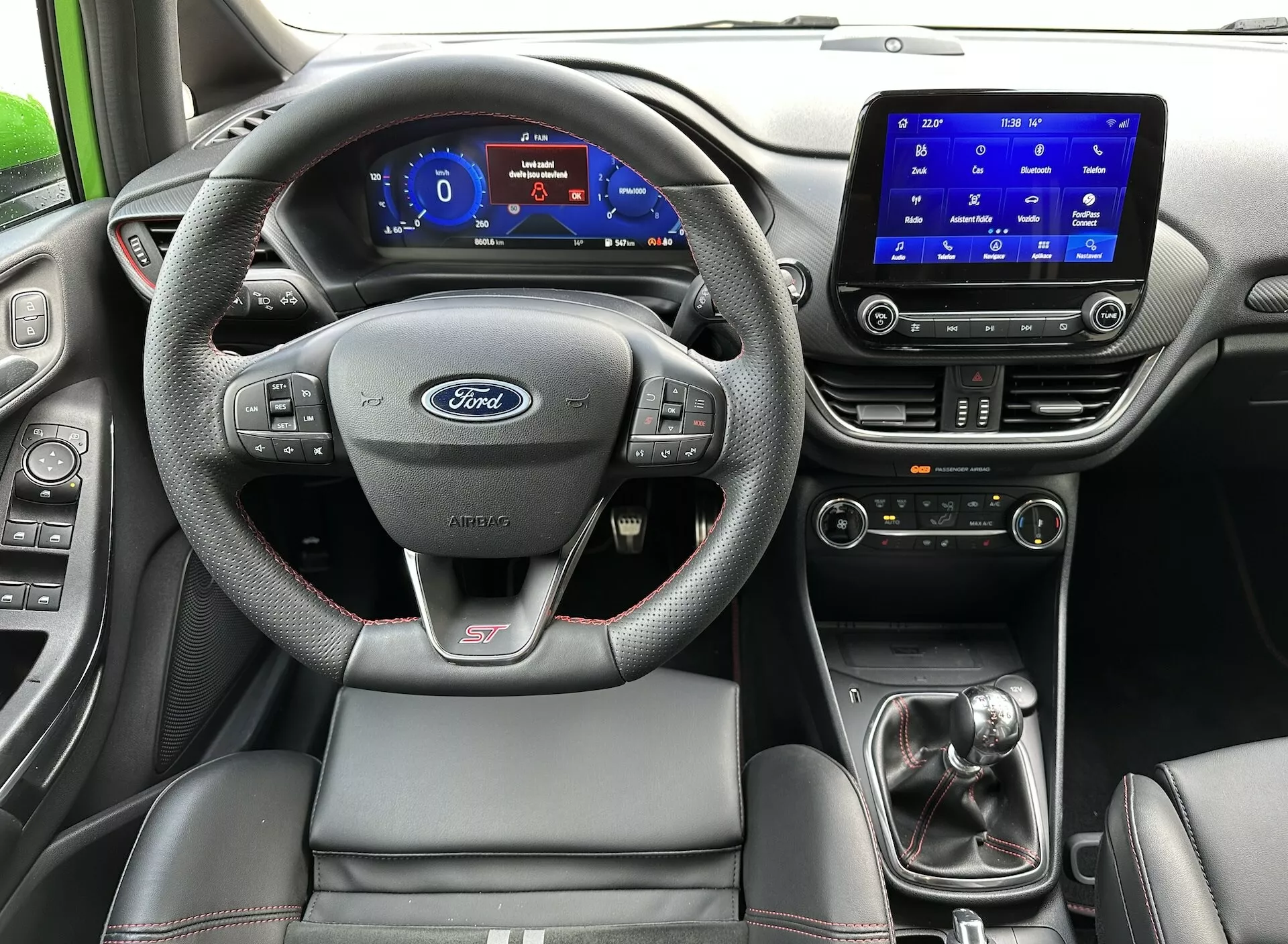 Interiörvy av Ford Fiesta 2022 från baksätet, med fokus på ratten med Ford-logotyp och ST-märke, digital instrumentpanel, pekskärmsinfotainmentsystem, manuell växelspak och sportigt förarsäte med kontrastsöm.