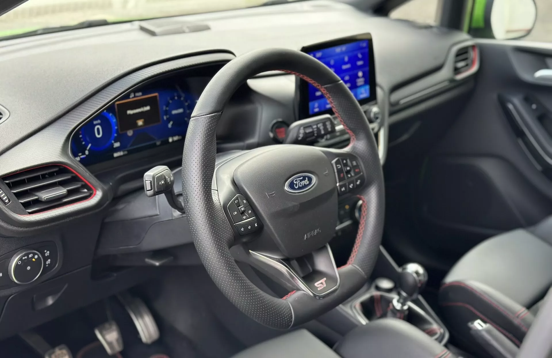 Interiör av Ford Fiesta 2022 från förarens perspektiv med läderratt med kontrollknappar, Ford-logotyp, digital instrumentpanel och modernt infotainmentsystem på instrumentbrädan