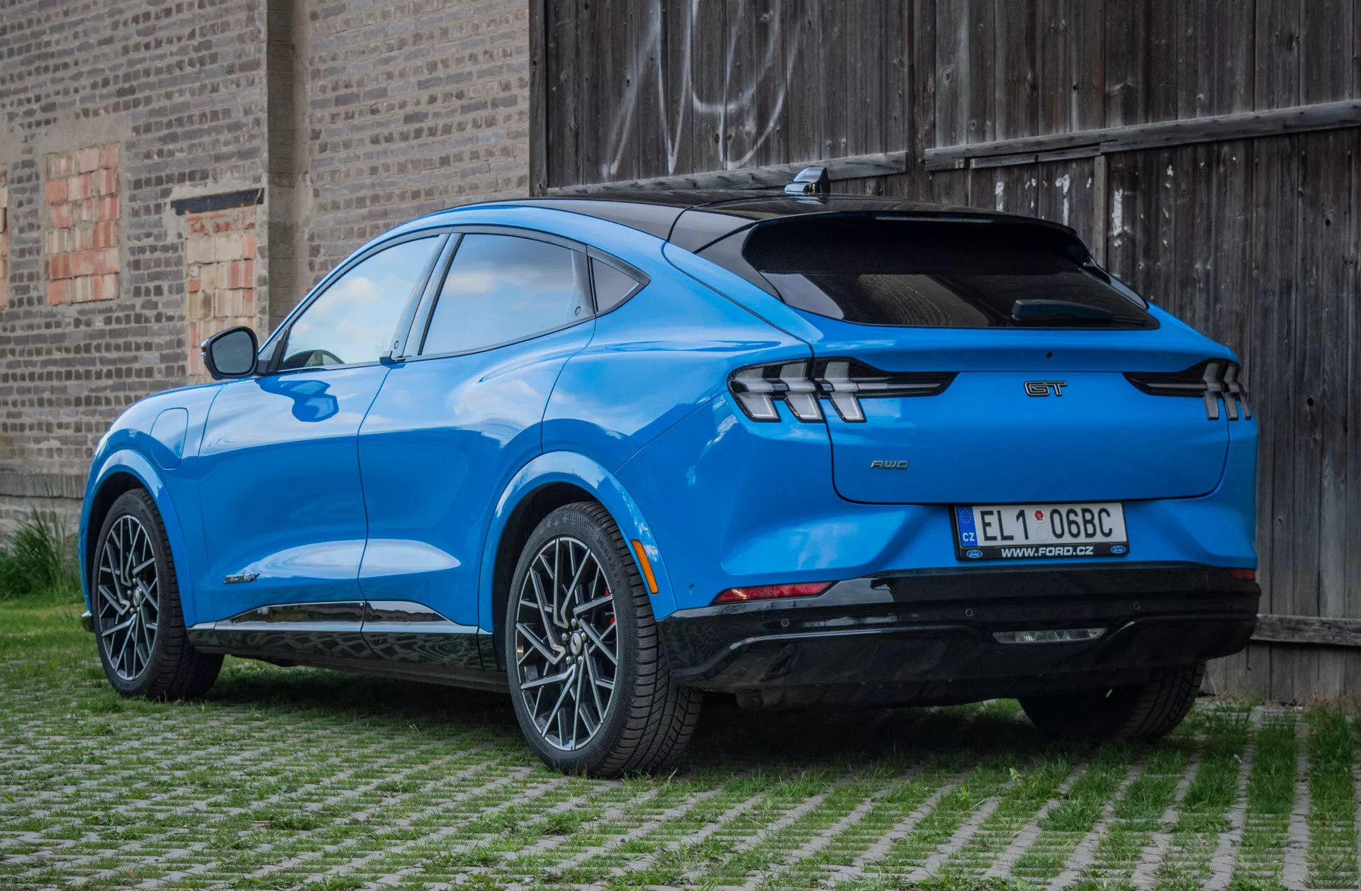 Ford Mustang Mach-E 2020 i klarblått, bakifrån tre fjärdedels vy som visar en slank design, legeringshjul, LED-bakljus och sportig spoiler, parkerad på ett gräsbeklätt område med murbakgrund