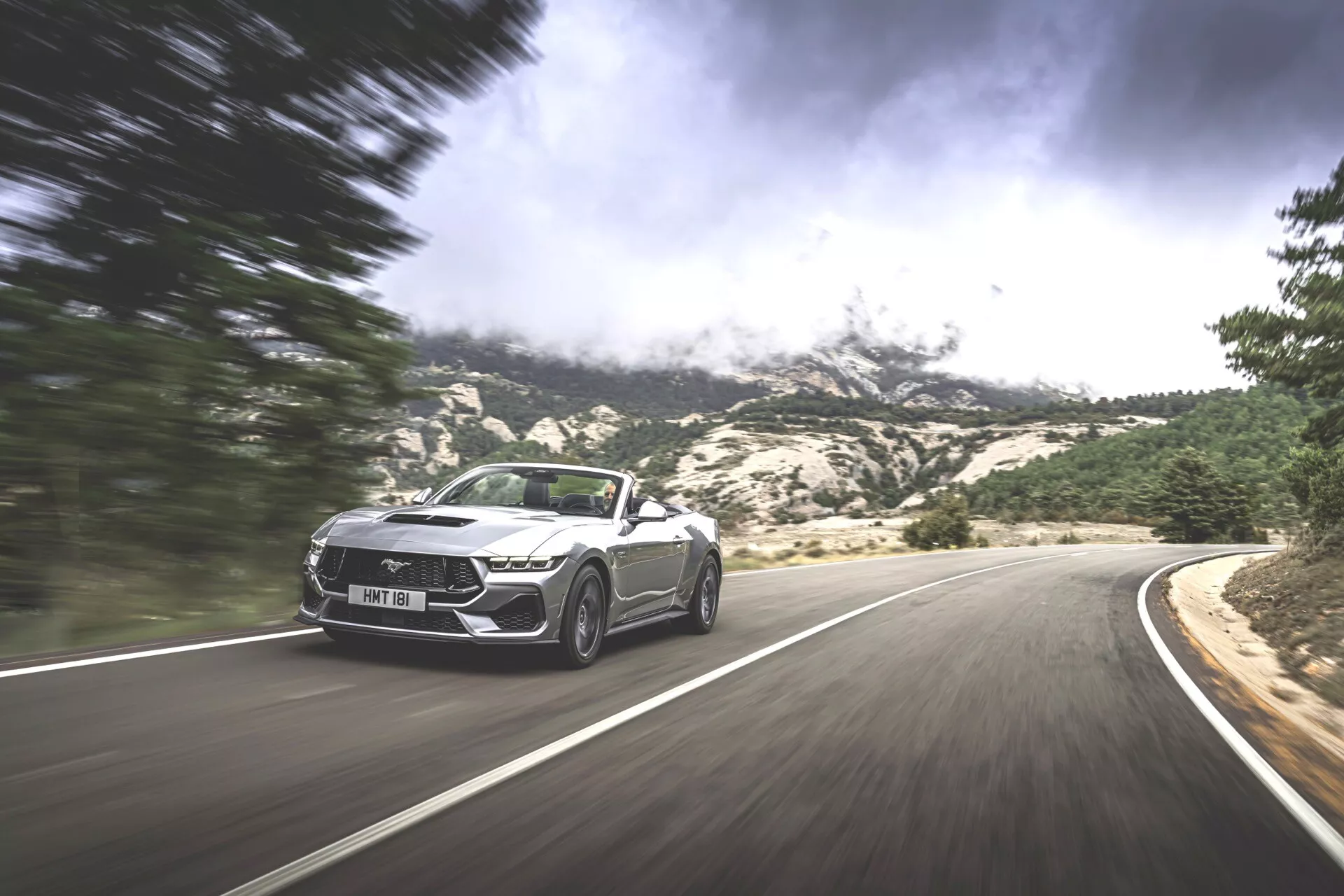Dynamisk bild av en Ford Mustang Cabriolet 2024 i rörelse på en slingrande bergsväg, som visar sportig design och lyxfunktioner mot en dramatisk bakgrund.