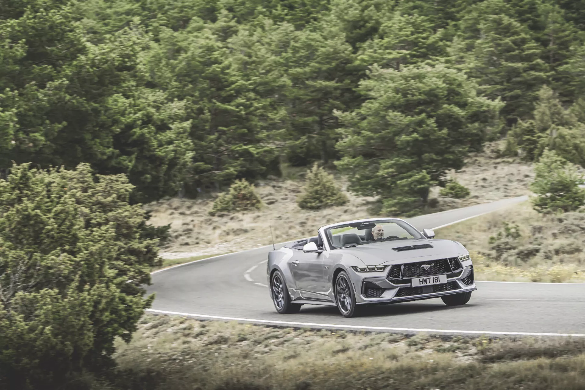 Ford Mustang Cabriolet 2024 i rörelse på slingrande väg, visar frontfasad, huva och sidoprofil med aerodynamisk design, LED-strålkastare och stora lättmetallhjul