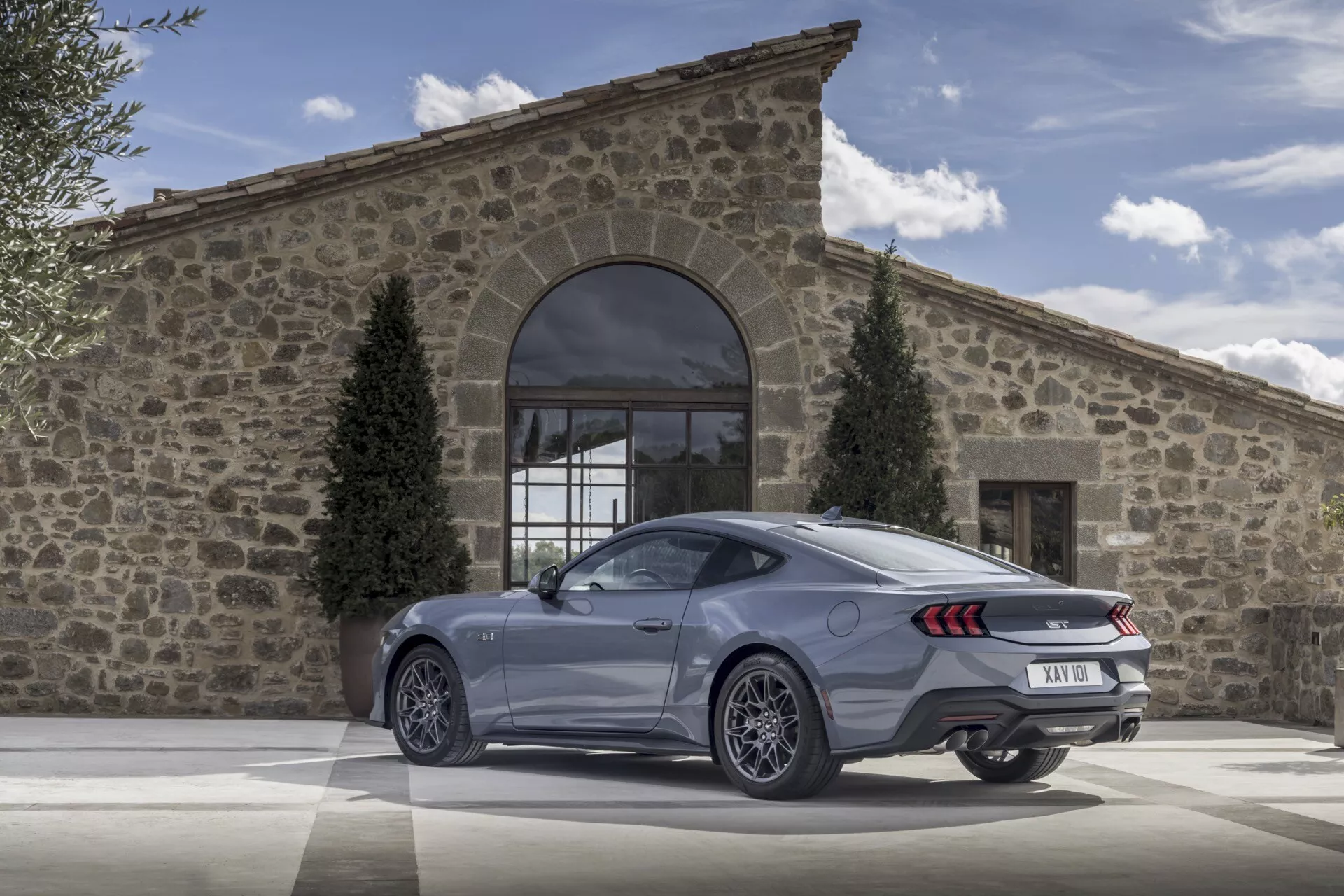 Ford Mustang coupé 2024 som visar bakifrån i trekvarts vy med LED-bakljus, dubbla avgasutblås och stora legeringshjul på en kullerstensbelagd uppfart framför en rustik byggnad på en klar dag, vilket betonar dess lyx- och prestandafunktioner.