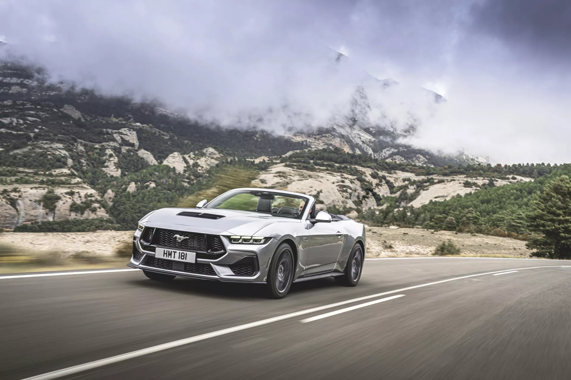 2024 Ford Mustang Cabriolet i rörelse, visar sina skarpa och vinklade designfunktioner, inklusive en framträdande grill med distinkt mönster, LED-strålkastare och skulpterad huva, sedd från en låg front-tre kvarts vinkel med en bergig bakgrund