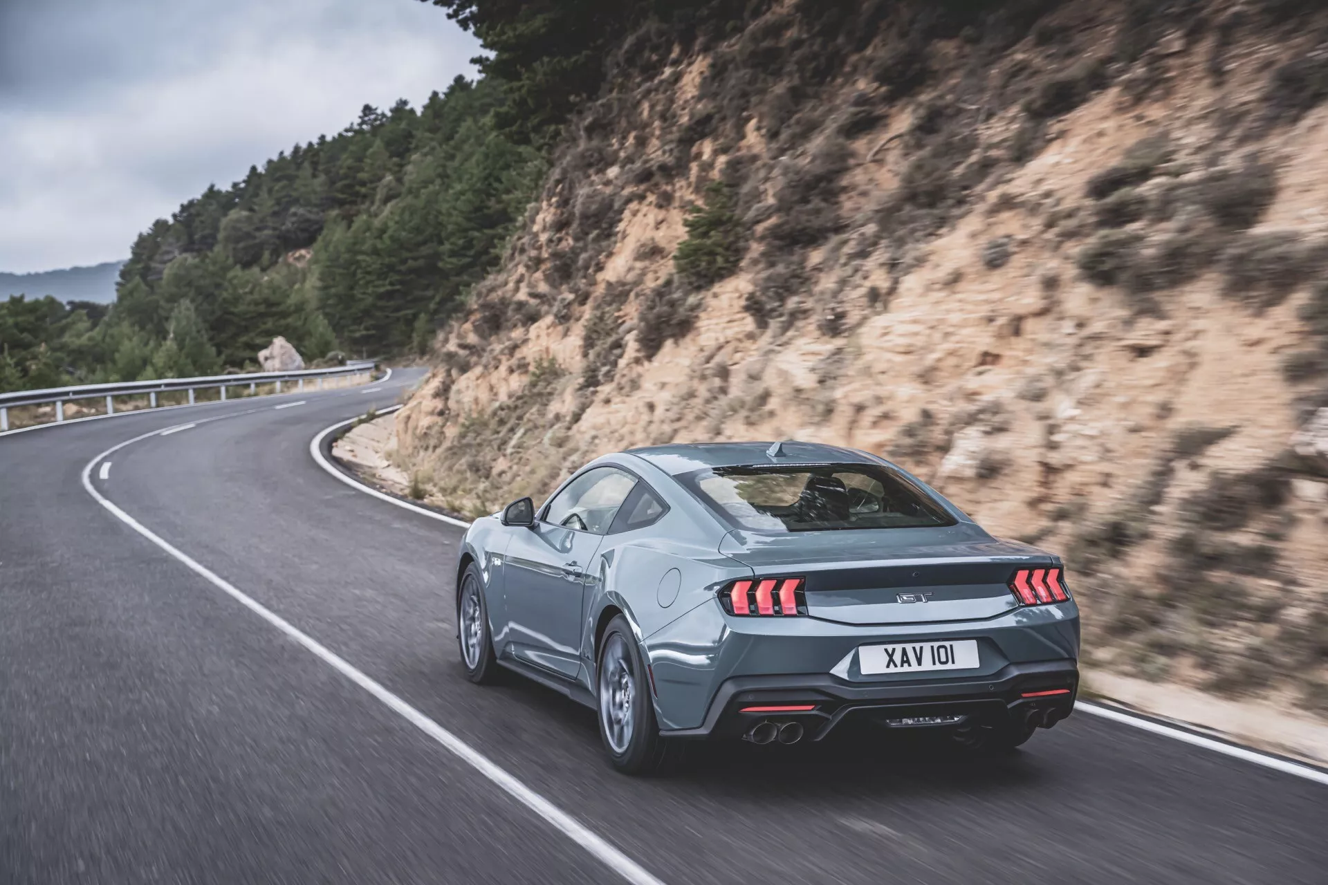 Silvercoupe Ford Mustang 2024 på slingrande väg: 3/4 bakvy som belyser LED-bakljus, dubbla avgasrör och aerodynamiska funktioner mot en naturskön bakgrund