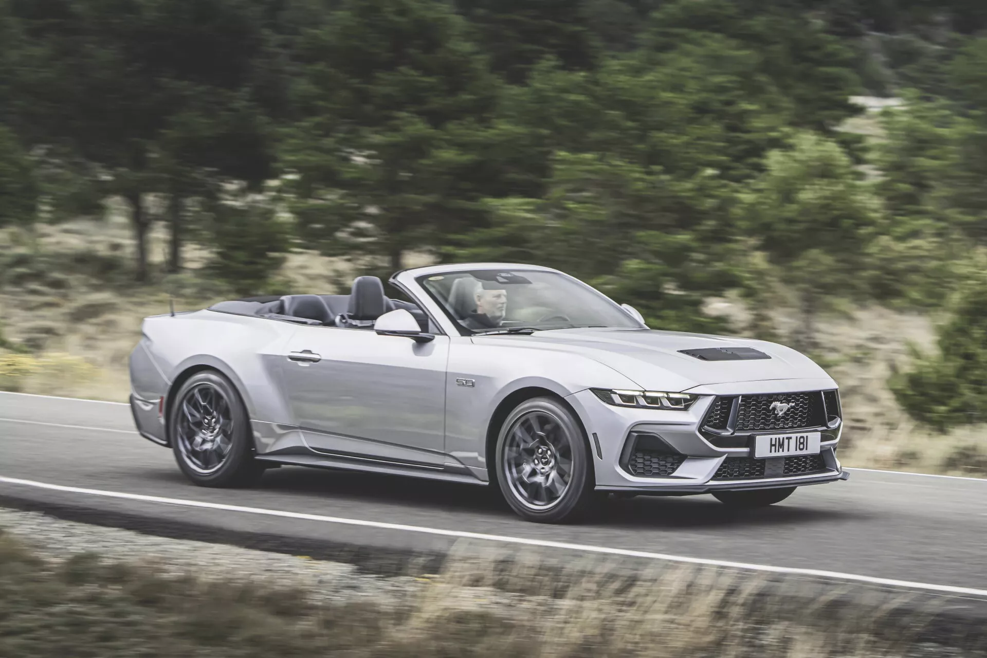 Ford Mustang Cabriolet 2024 i silver, som visar sin aggressiva styling och avancerade funktioner i rörelse på en motorväg, fångad från förarsidan i ett tre-fjärdedels perspektiv