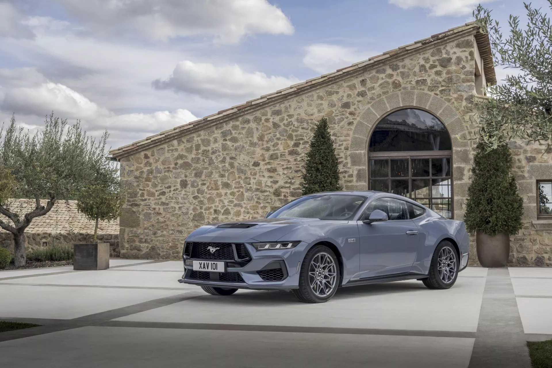 Ford Mustang 2024 i snygg metallfinish parkerad framför en exklusiv byggnad med stenfasad, som visar upp sin sportiga och eleganta design med en detaljerad vy av dess vinklade LED-strålkastare, distinkt frontgaller och flerspakiga legeringshjul