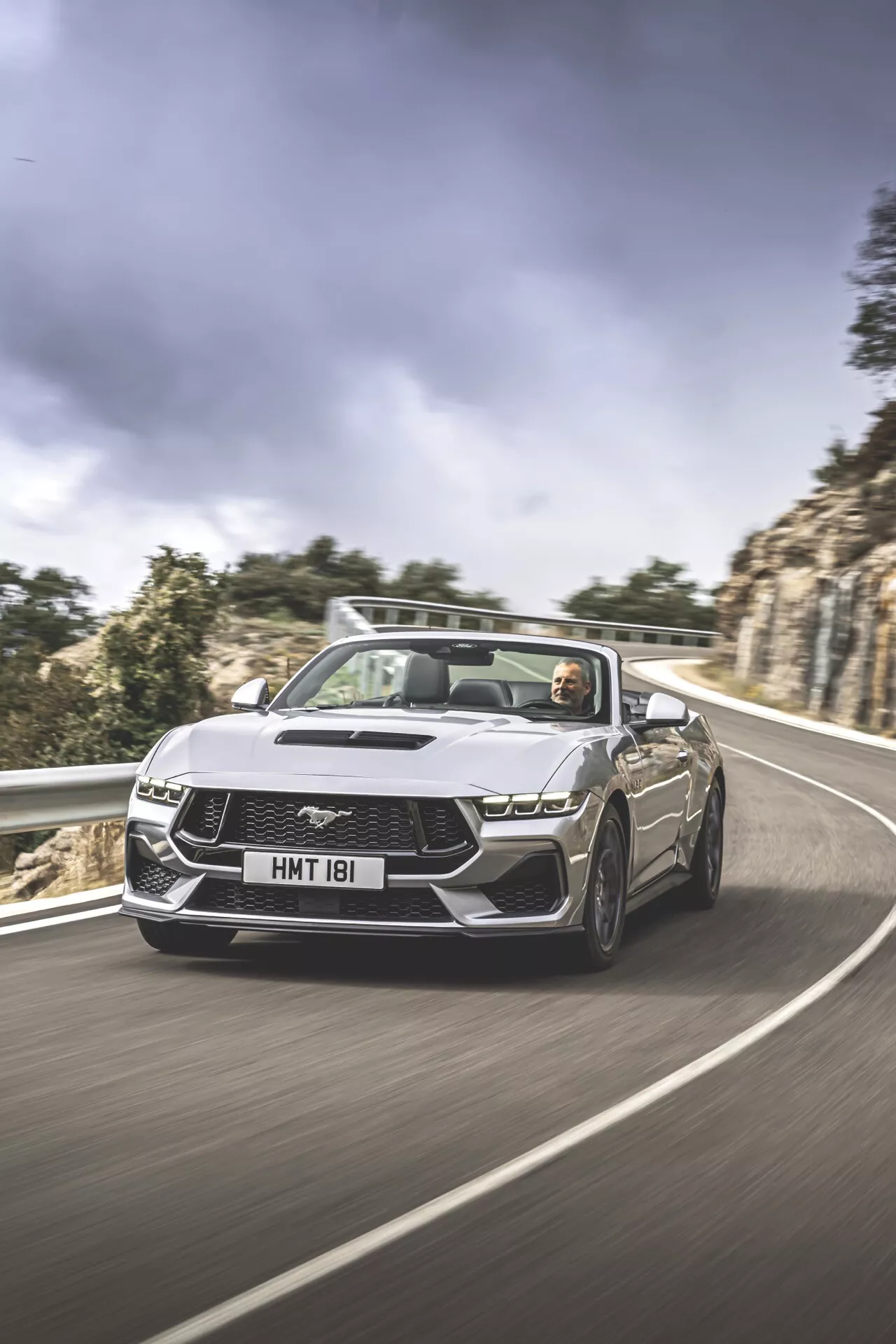 2024 Ford Mustang 2:a generationens cabriolet i dynamisk körning på en slingrande väg som visar LED-strålkastare, emblem grill, nedfällbart cabriolettak med synlig interiör och förare, designad för prestanda och lyx