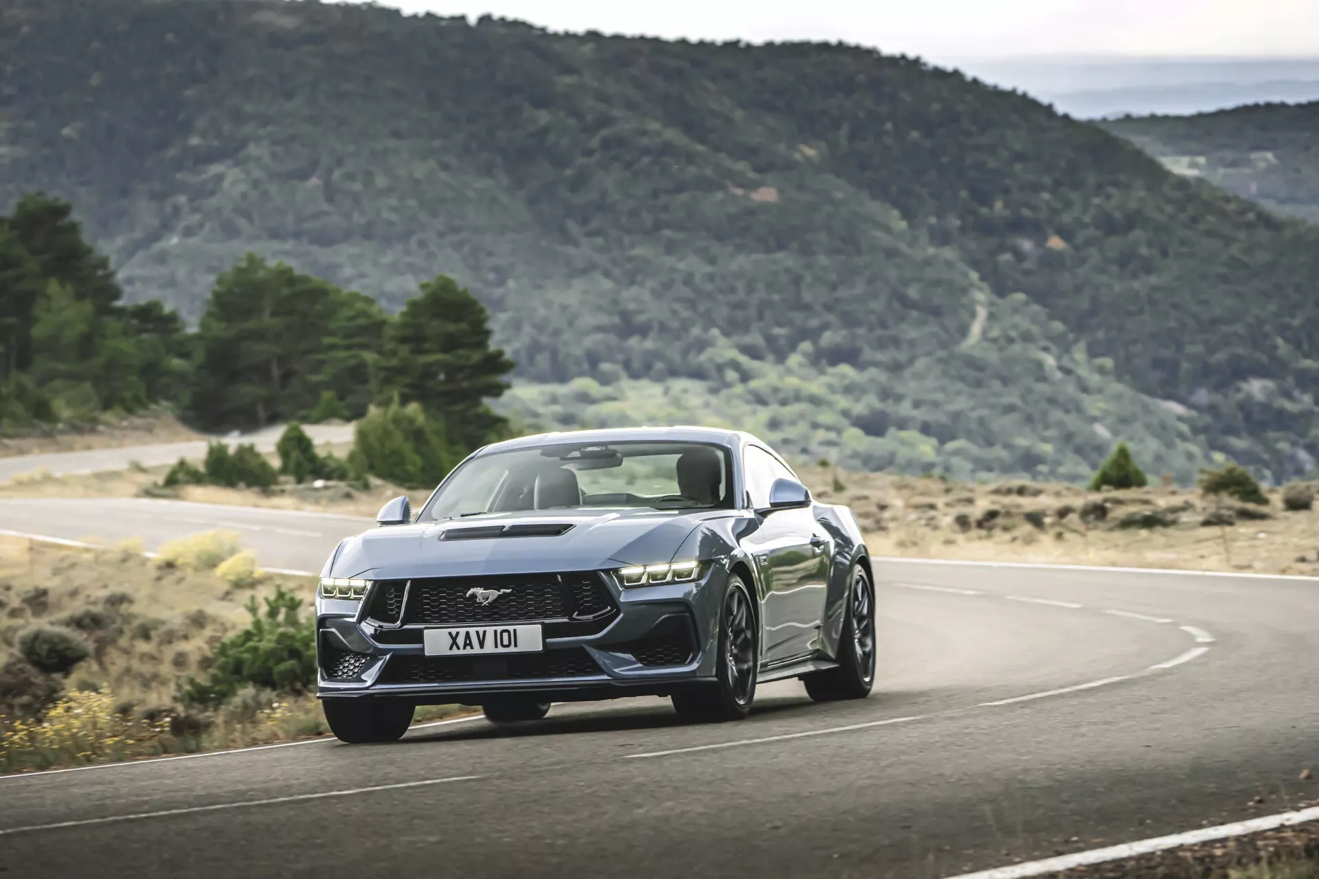 Ford Mustang andra generationen 2024 i en dynamisk körscen på en slingrande bergsväg, som visar en aggressiv hållning, moderna LED-strålkastare, intrikat grill, muskulösa huva linjer och sportiga legering hjul