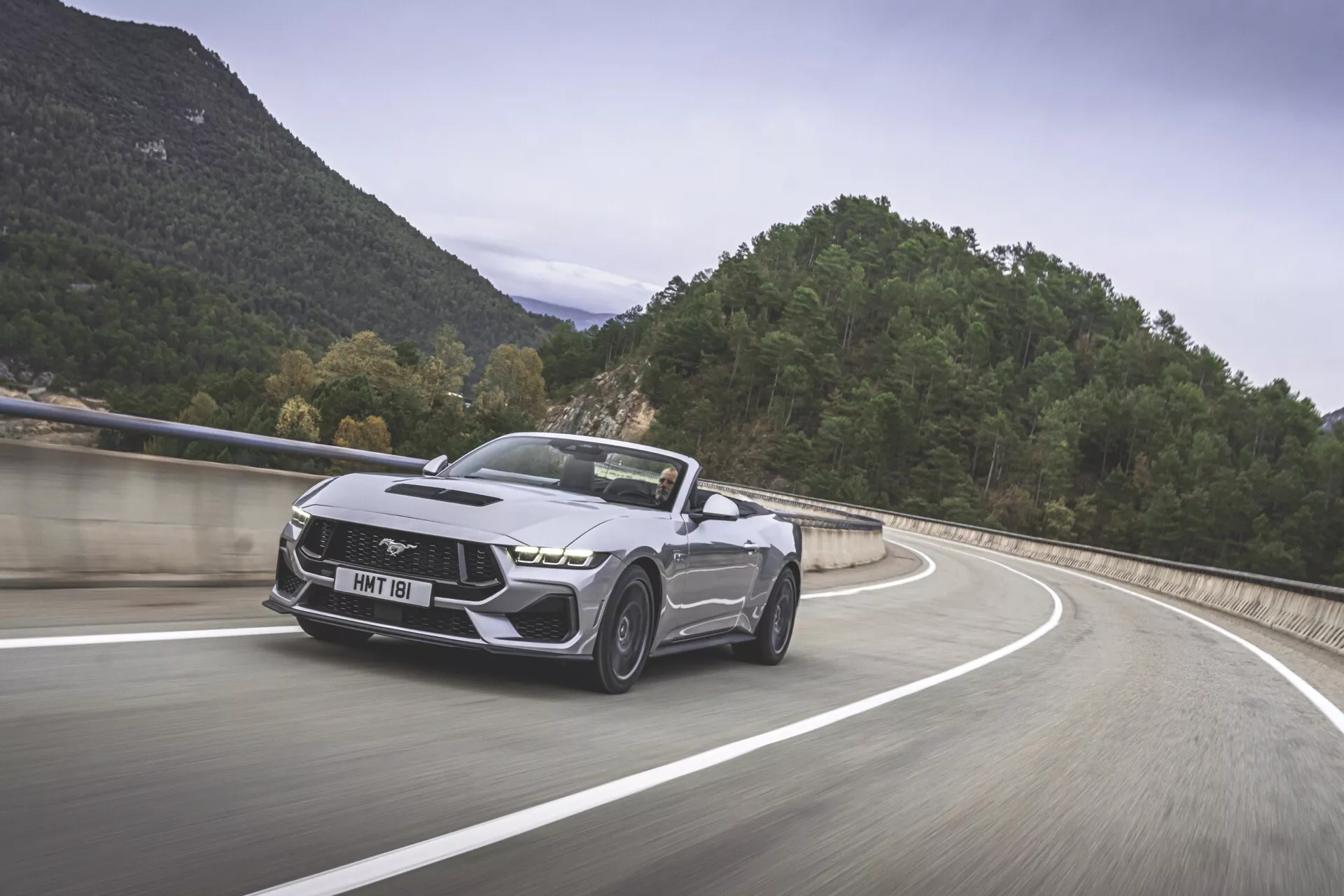 Ford Mustang 2024 i rörelse på en bergig väg, som visar upp sin moderna muskelbilsdesign med en ikonisk galopperande hästemblem, aggressiv främre del med eleganta strålkastare, sportig stötfångare, skulpterade huvar, distinkta legeringshjul och cabriolet-topp i metallisk finish.