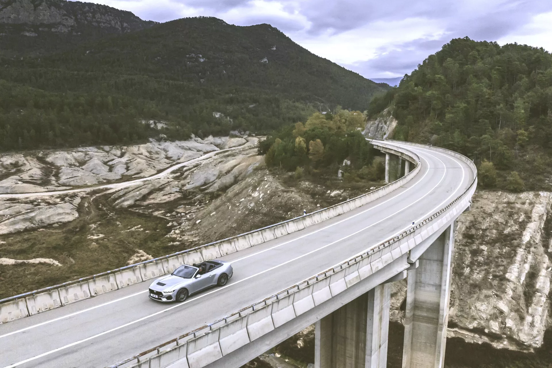 Ford Mustang 2024 som färdas på en bro i en kuperad landskap, fångad från en hög vinkel som visar den främre vänstra sidan och översikten med fokus på den metalliska färgen och designfunktioner som grillen fram och legeringshjulen under naturligt ljus