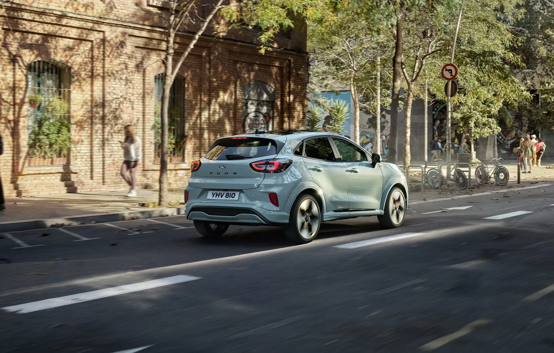 Ford Puma 2024 (II-facelift) ljusfärgad halvkombi i rörelse på en stadsgata från en tre fjärdedels bakre vinkel som visar bakljus, bakfönster, sidodörrar och lättmetallfälgar
