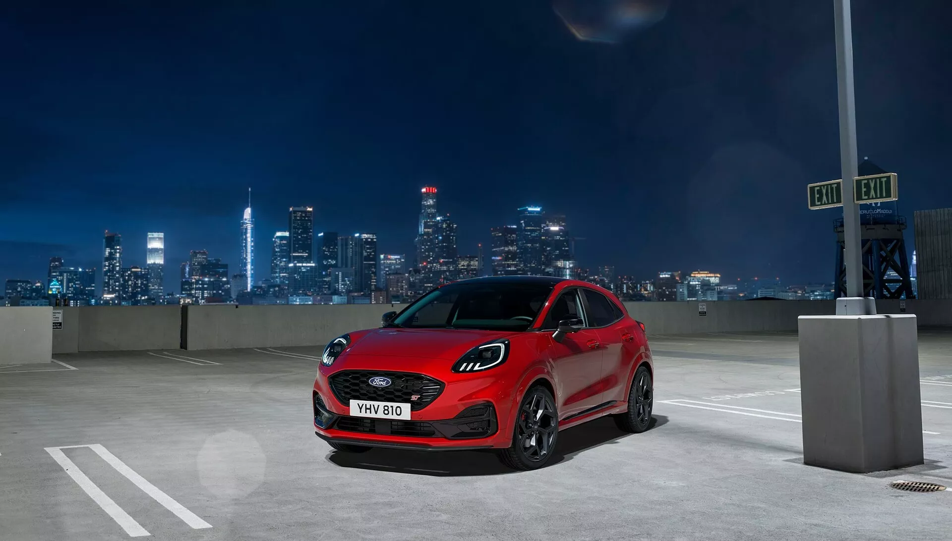 2024 Ford Puma (II facelift) röd hatchback parkerad på en utomhusparkering på natten med upplyst stadssilhuett i bakgrunden, visar aggressiva LED-strålkastare, stor grill och sportig profil från en låg vinkel tre fjärdedels framsida